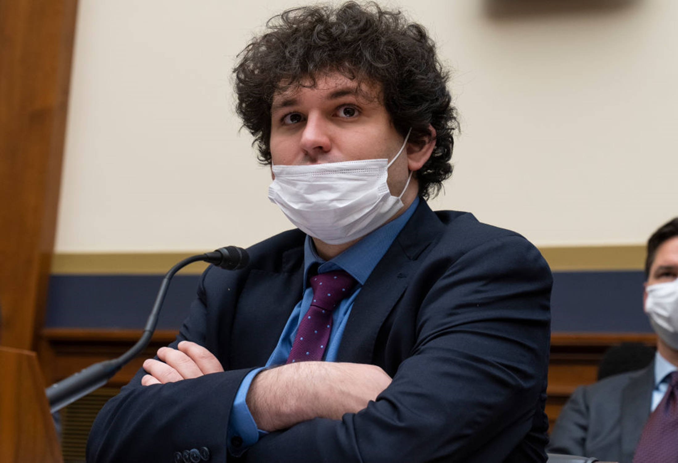 桌布 壁紙 — 一名留著捲髮、戴著白色口罩、身穿西裝的男子，在正式聆訊室內交叉雙臂坐著。
