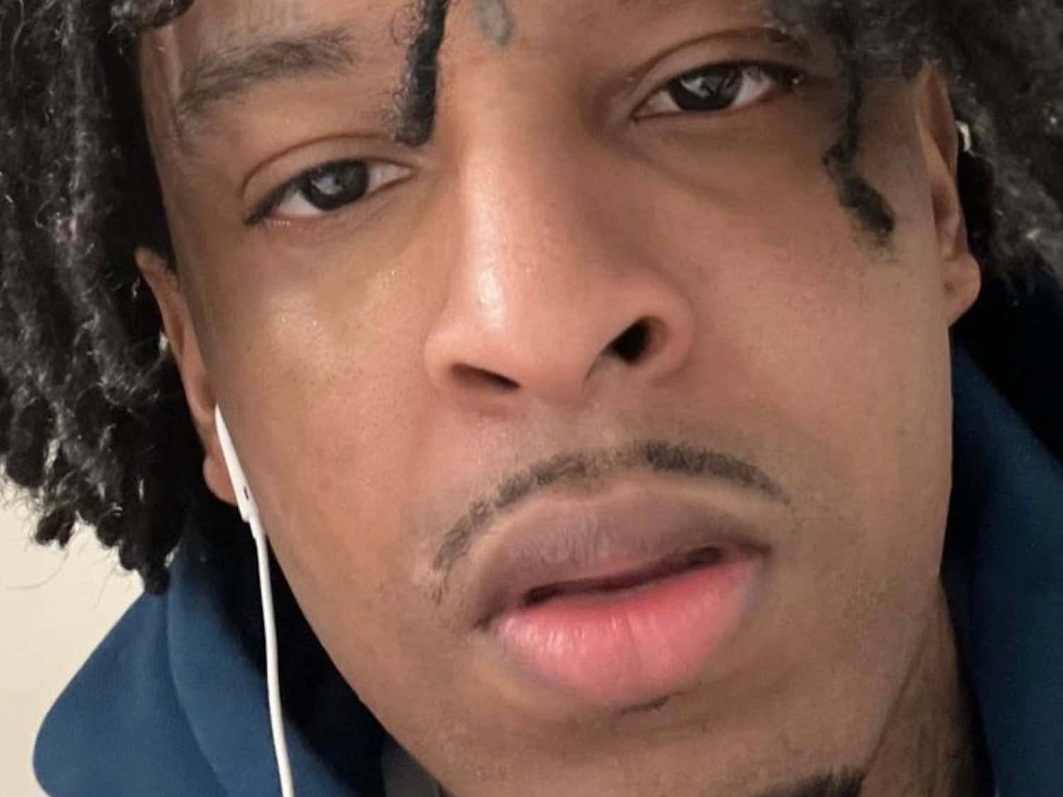 21 Savage 桌布 壁紙 — 一名留著捲髮、臉上有刺青的男子特寫肖像，戴著白色耳機和藍色連帽衫。