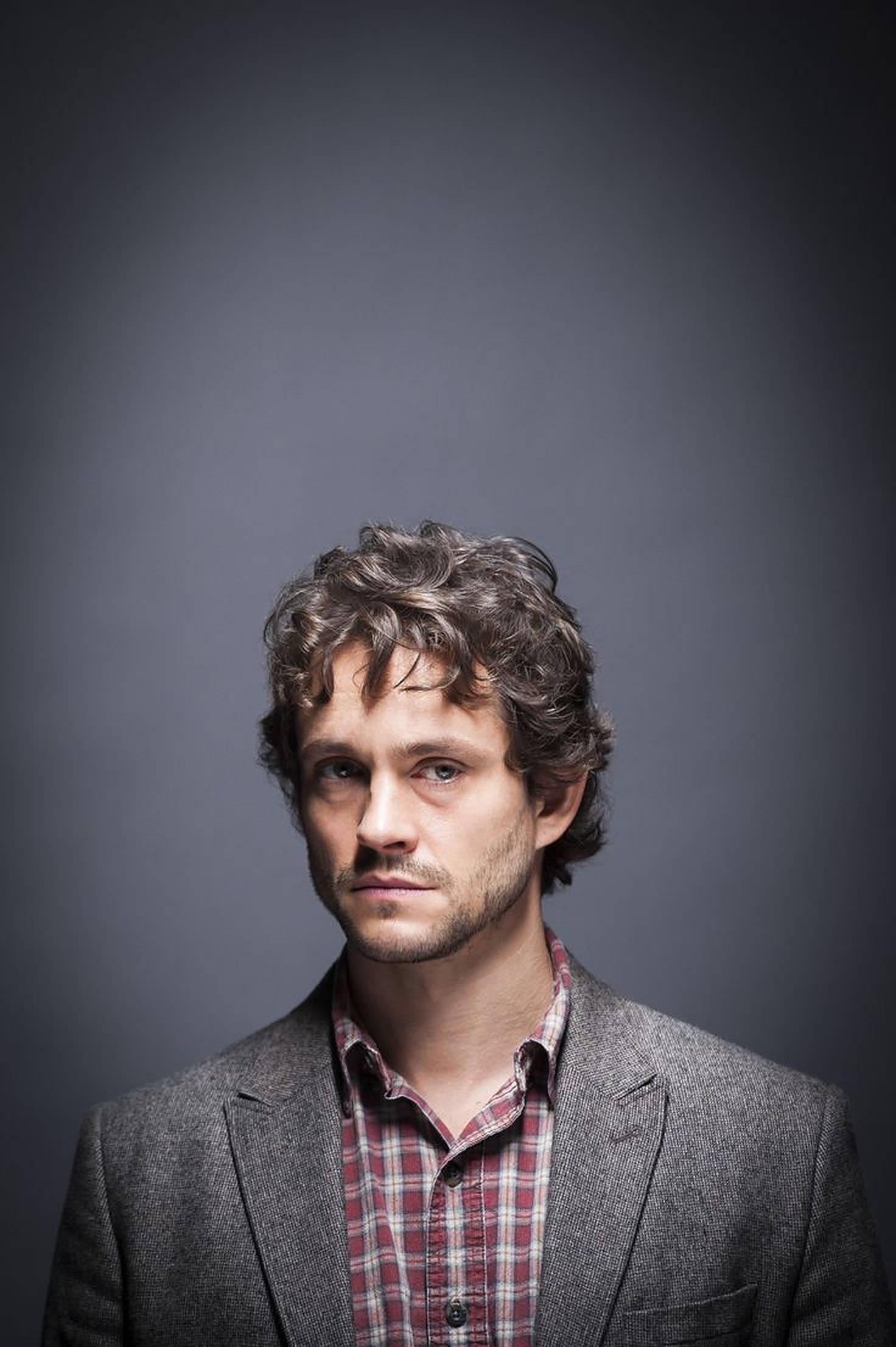 Hugh Dancy 桌布 壁紙 — 一名留著捲髮和鬍鬚的男子，在深灰色攝影棚背景前，穿著灰色西裝外套內搭格紋襯衫。