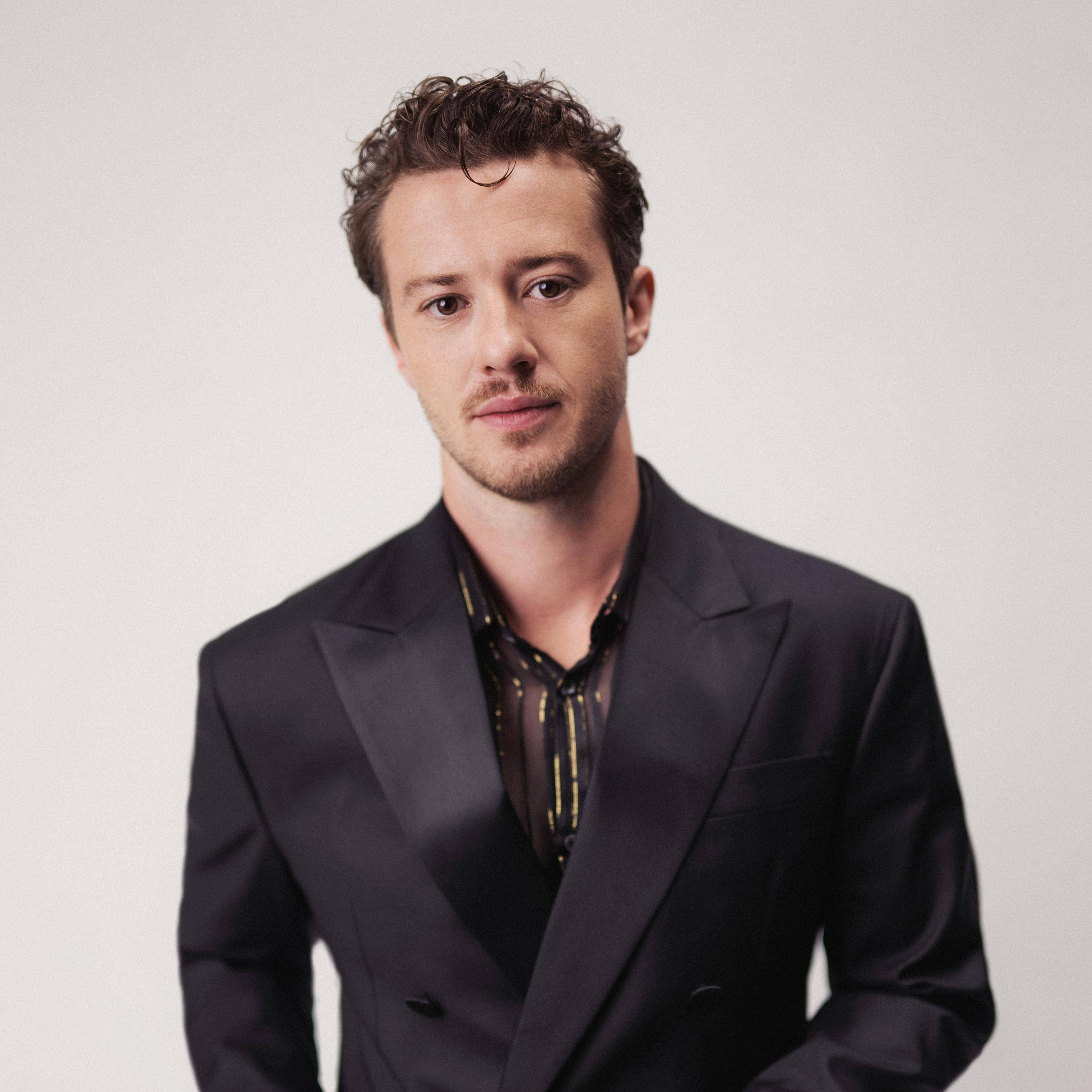Celebrity 桌布 壁紙 — 一名留著捲髮和鬍鬚的男子，身穿黑色西裝，內搭圖案襯衫，背景為純白色。