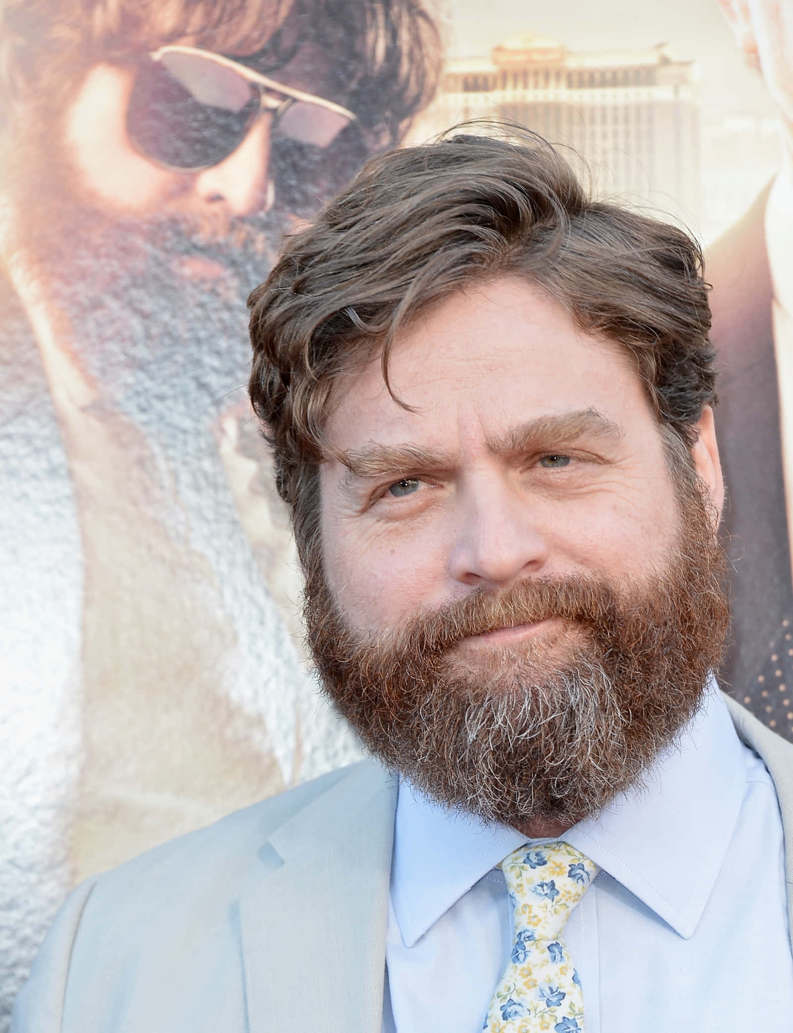 Zach Galifianakis 桌布 壁紙 — 一名留著濃密鬍鬚和波浪髮型的男士，穿著淺色西裝和花卉領帶，在電影海報背景下微笑。