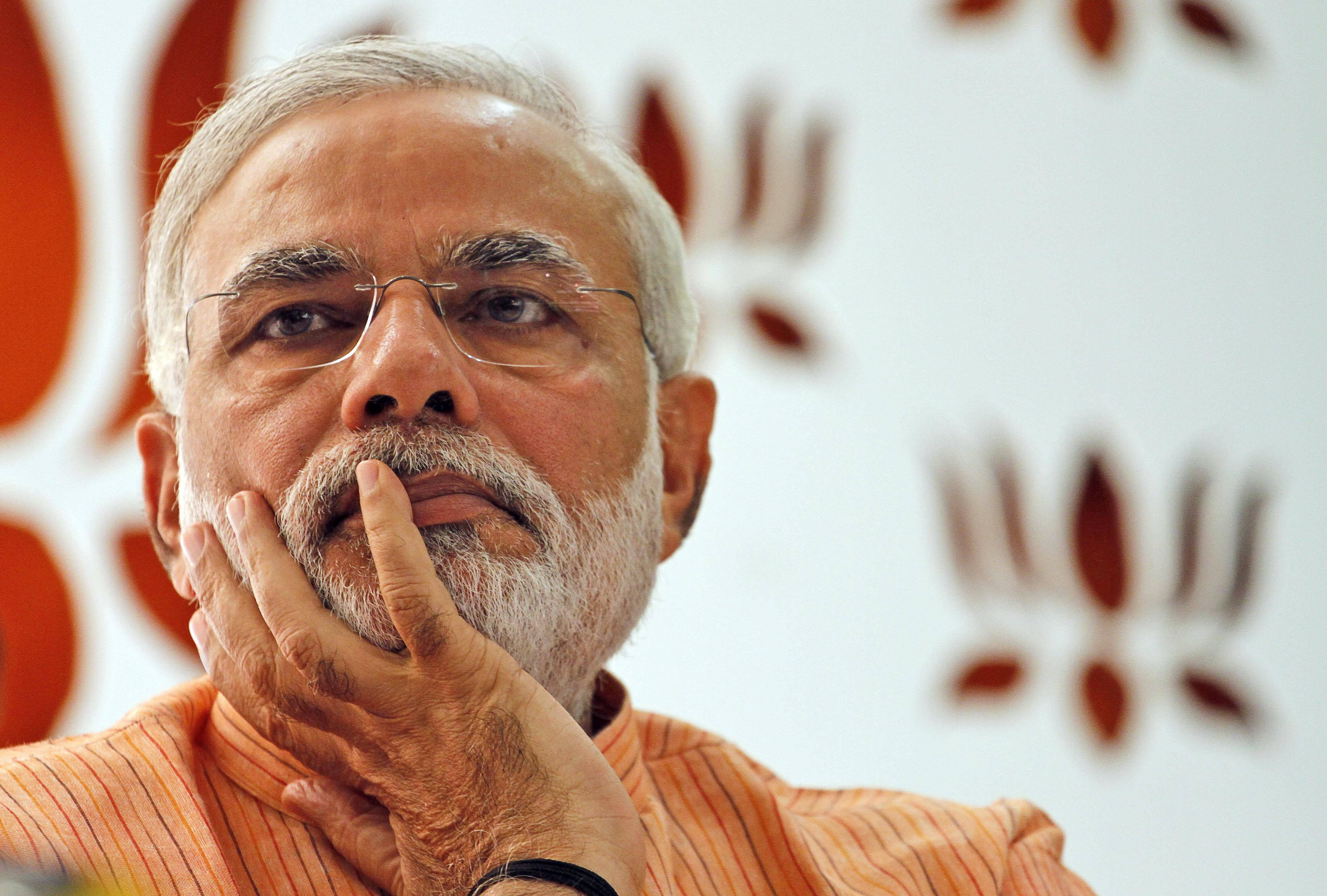 Narendra Modi 桌布 壁紙 — 一名留著白鬍子、戴眼鏡的男子，在模糊的橙色和白色背景前，將下巴擱在手上的特寫肖像。