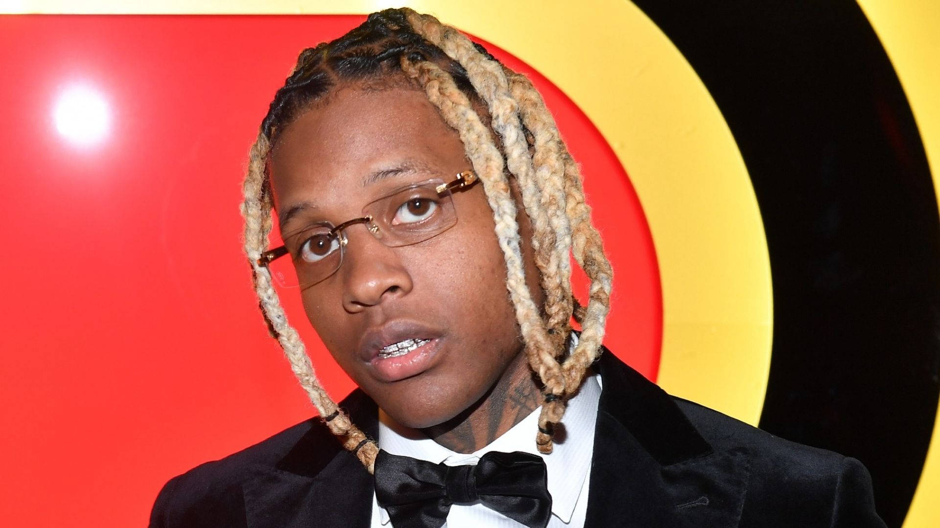 Lil Durk 桌布 壁紙 — 一名留著辮子、戴眼鏡的男子，在搶眼的紅、黃、黑圓形背景前穿著黑色燕尾服。