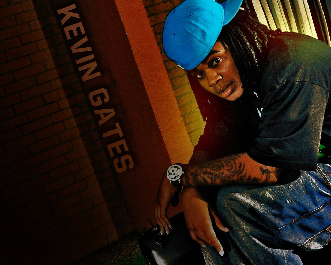 Kevin Gates 桌布 壁紙 — 一名留著辮子、戴藍色帽子的男人靠在一面磚牆上，牆上垂直寫著「KEVIN GATES」，他露出有刺青的手臂，穿著牛仔布。