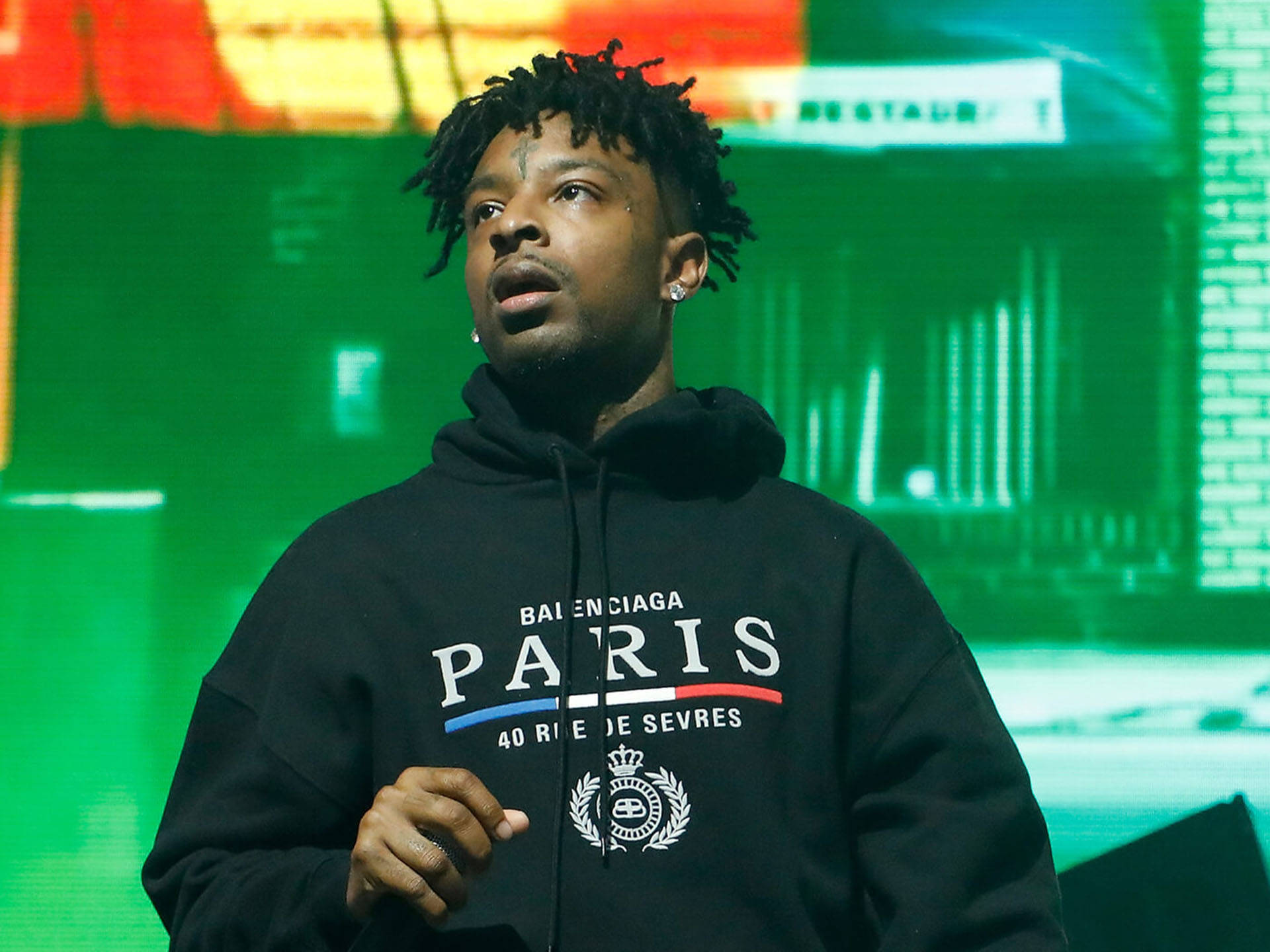 savage 桌布 壁紙 — 一名留著辮子、身穿黑色 Balenciaga Paris 連帽衫的男子站在舞台上，舞台被鮮豔的綠色和紅色燈光照亮。