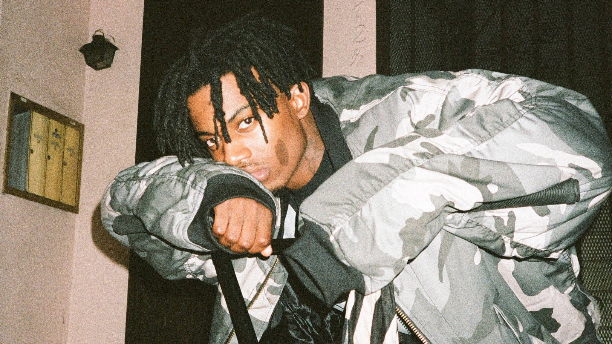 Carti 桌布 壁紙 — 一名留著辮子的年輕男子身穿迷彩外套，在光線昏暗的室內空間向前傾，專注地凝視鏡頭。