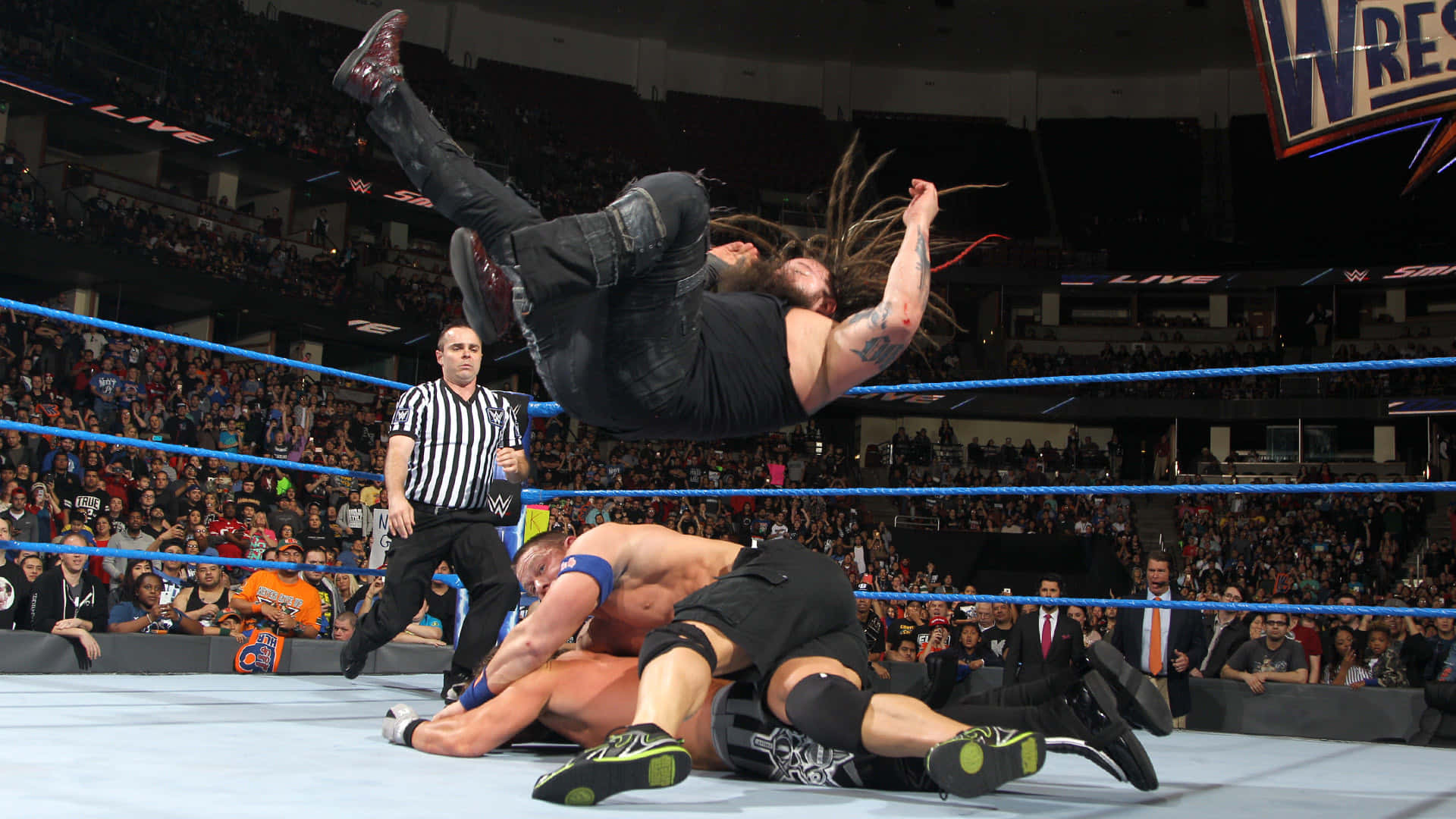 WWE 桌布 壁紙 — 一名留著辮子的摔角手在半空中跳向一名被壓在擂台上的對手，場館內座無虛席。