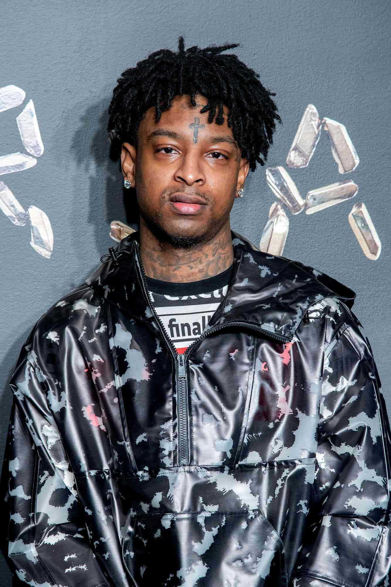 21 Savage 桌布 壁紙 — 一名留著辮子頭、帶有十字架刺青的男子，身穿閃亮迷彩外套，凝視前方，背景是帶有水晶貼紙的灰色牆壁。