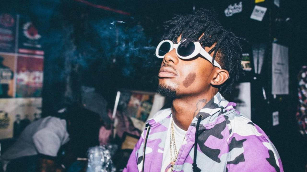 Carti 桌布 壁紙 — 一名留著辮子頭、戴白色太陽眼鏡的男子，在昏暗的房間裡吐煙，牆上貼著海報。