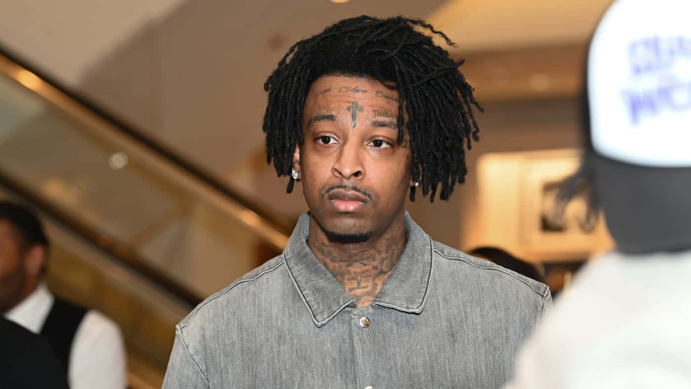 21 Savage 桌布 壁紙 — 一名留著辮子頭、臉上有刺青的男子，在溫暖照明的室內環境中，身後有電扶梯，他專注地凝視著。