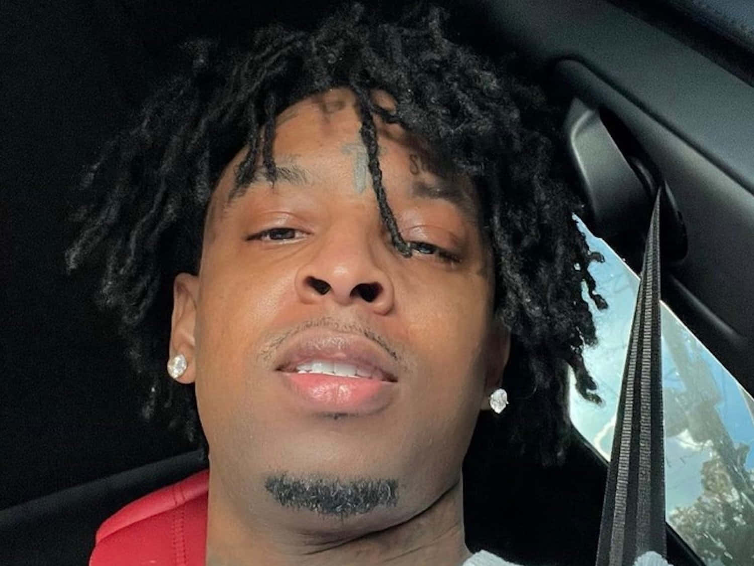 21 Savage 桌布 壁紙 — 一名留著辮子頭、臉上有刺青的男子，身穿紅色襯衫坐在車內的近距離特寫。