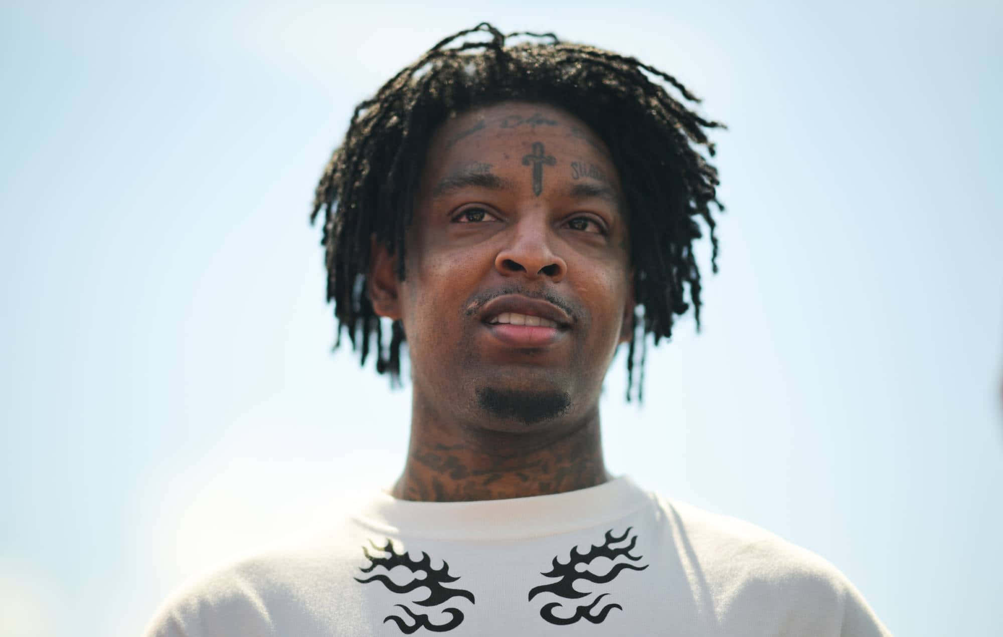 21 Savage 桌布 壁紙 — 一名留著辮子頭、臉上有刺青的男子，身穿帶有黑色火焰圖案的白襯衫，仰望明亮的天空。