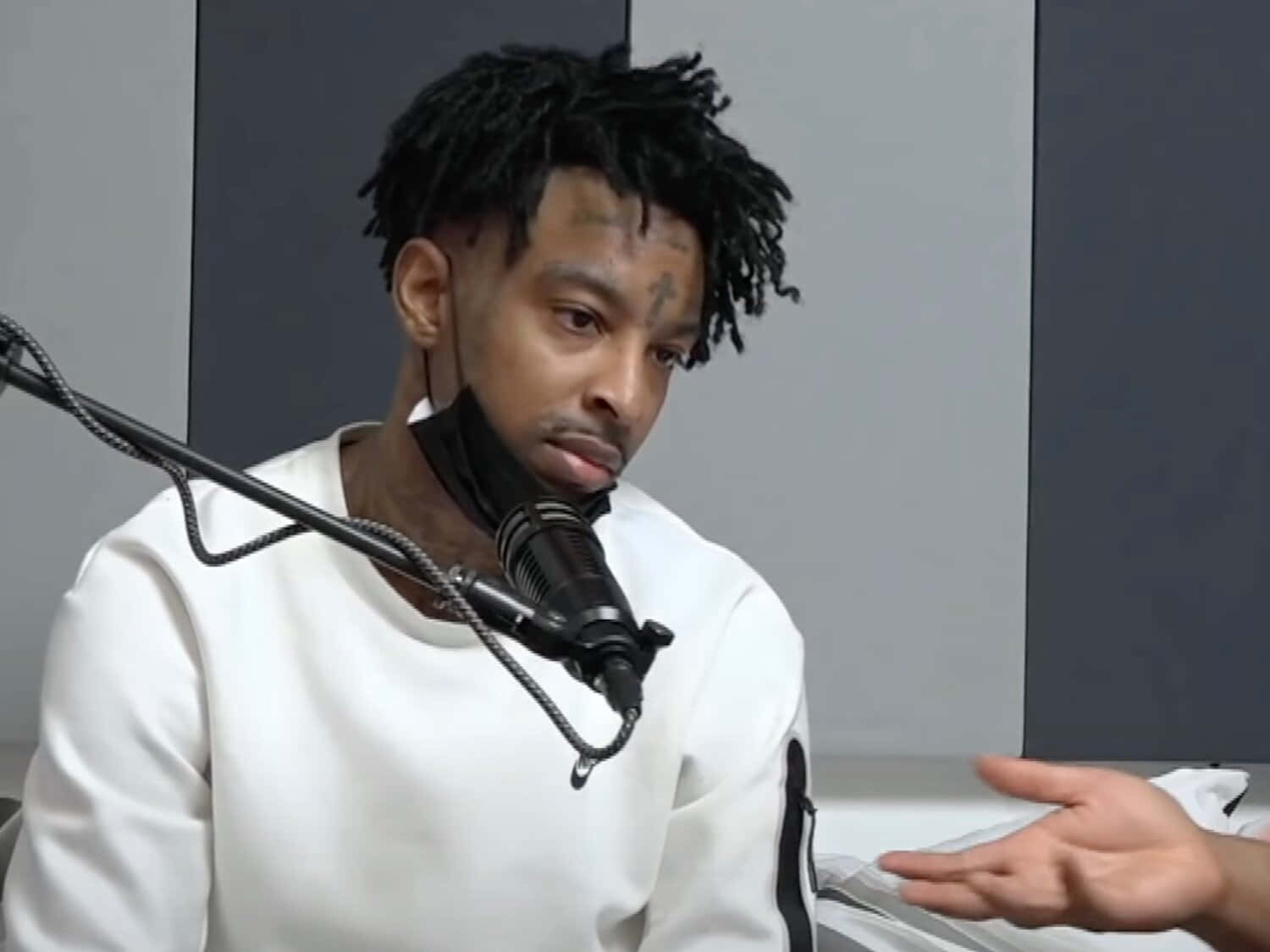 21 Savage 桌布 壁紙 — 一名留著辮子頭、臉上有刺青的男子，身穿白色連帽上衣，坐在錄音室裡對著麥克風說話。