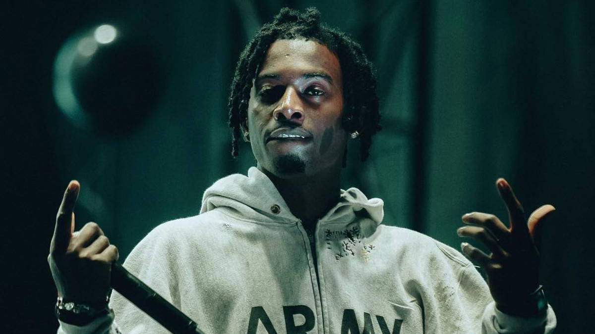 Carti 桌布 壁紙 — 一名留著辮子頭、身穿白色連帽衫的男子，在昏暗的綠色舞台燈光下，一邊拿著麥克風一邊做手勢。
