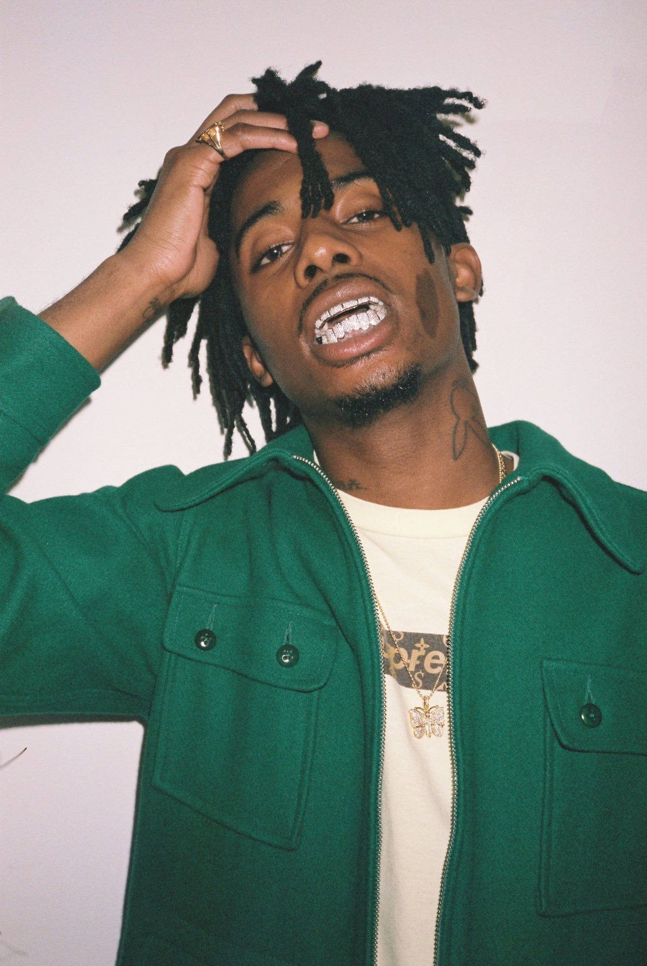 playboi carti pfp 桌布 壁紙 — 一名留著辮子頭、身穿綠色外套的男子，將頭靠向純色背景，露出燦爛的笑容。