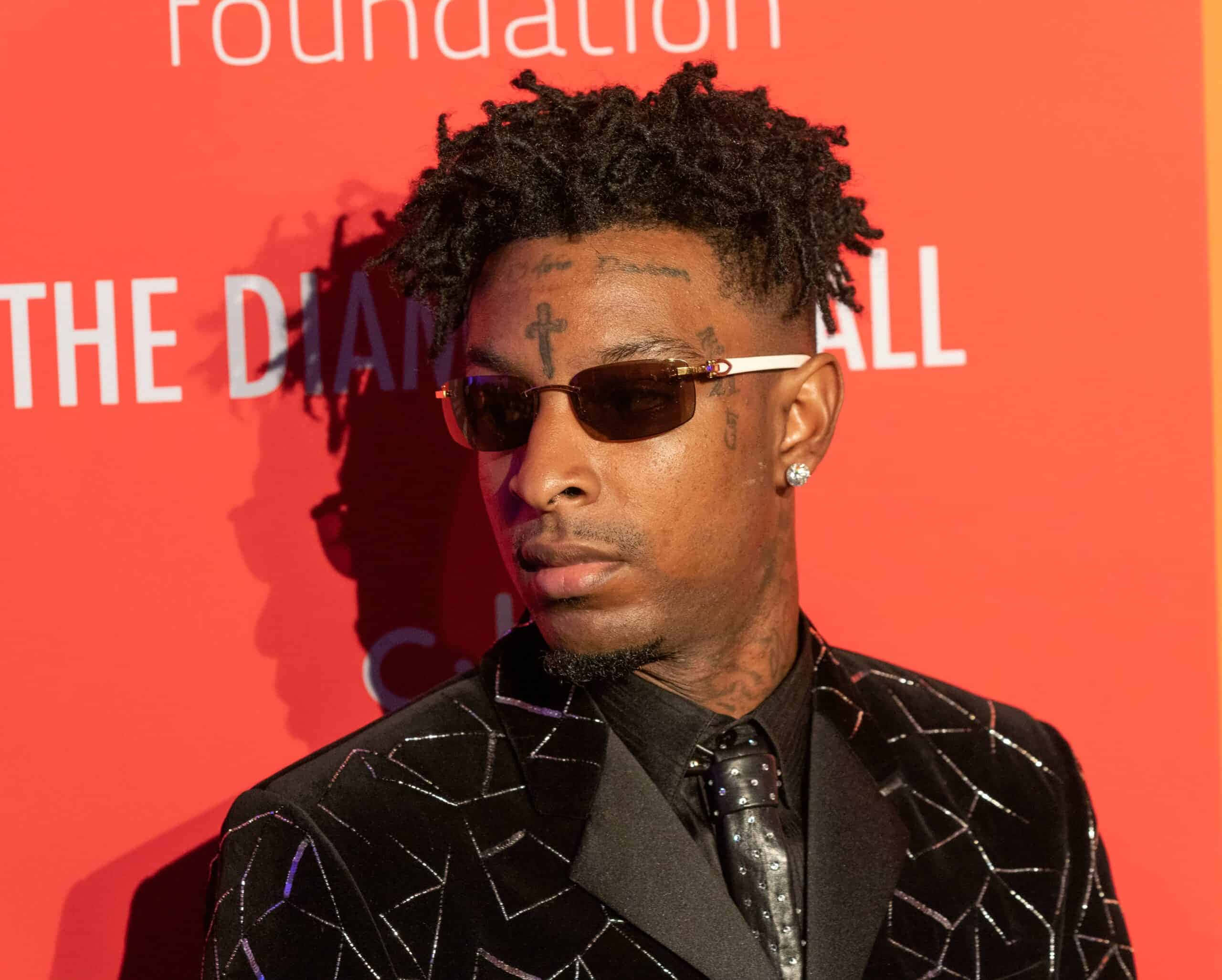 21 Savage 桌布 壁紙 — 一名留著辮子頭和刺青的男子，戴著太陽眼鏡，穿著閃亮的黑色西裝，背景是紅色活動背景。