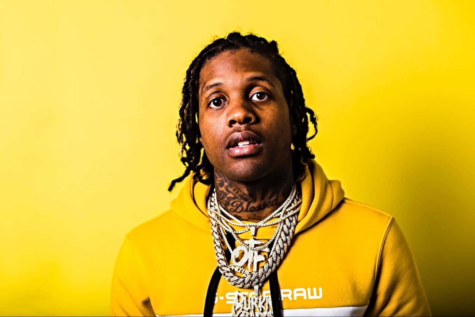 Lil Durk 桌布 壁紙 — 一名留著辮子頭和刺青的男子，穿著黃色連帽衫和層疊的金鏈，背景是純黃色。