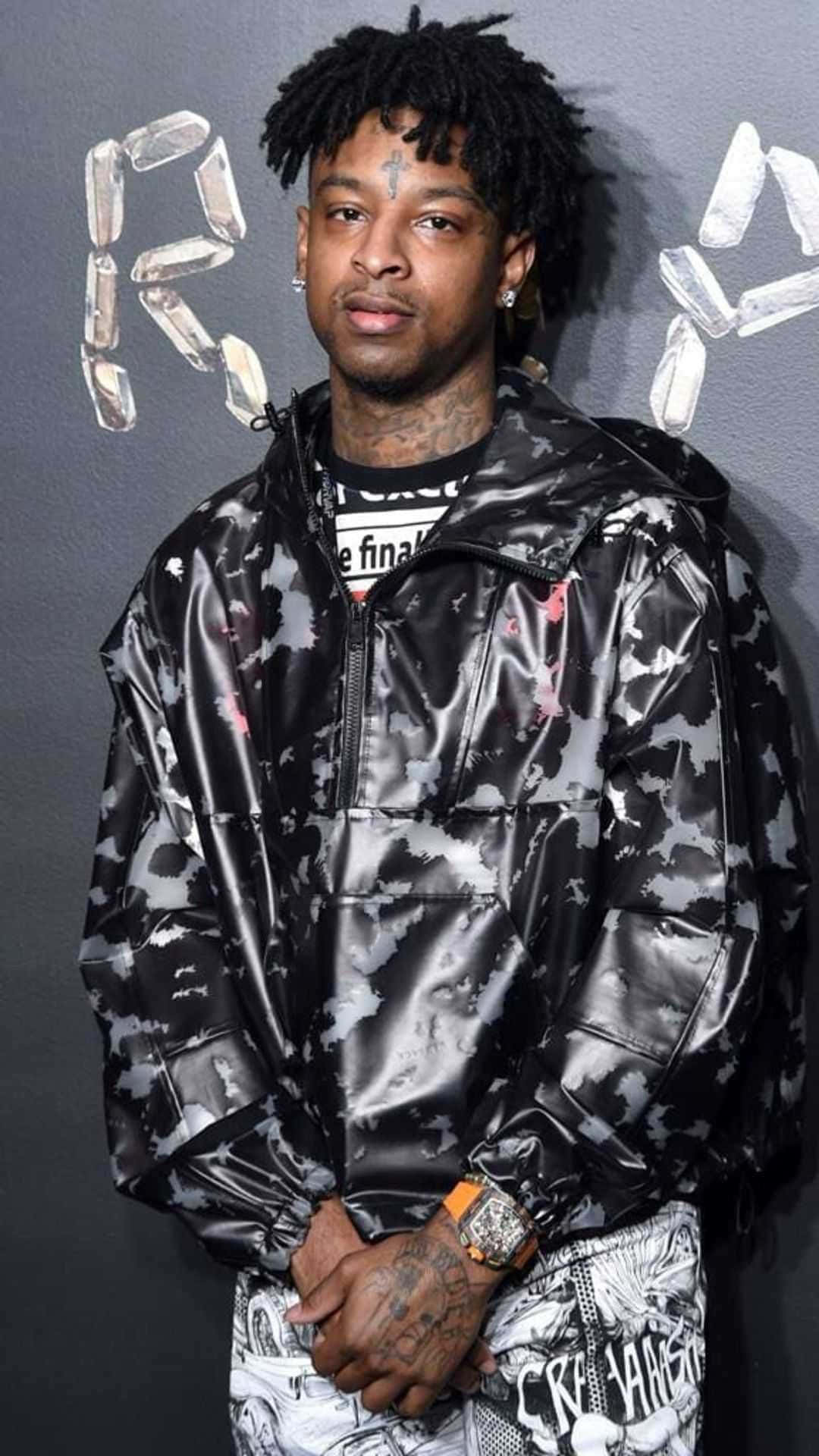 21 Savage 桌布 壁紙 — 一名留著辮子頭和刺青的男子，穿著閃亮的黑白迷彩外套，背景是灰色牆壁，上面有水晶般的字母。