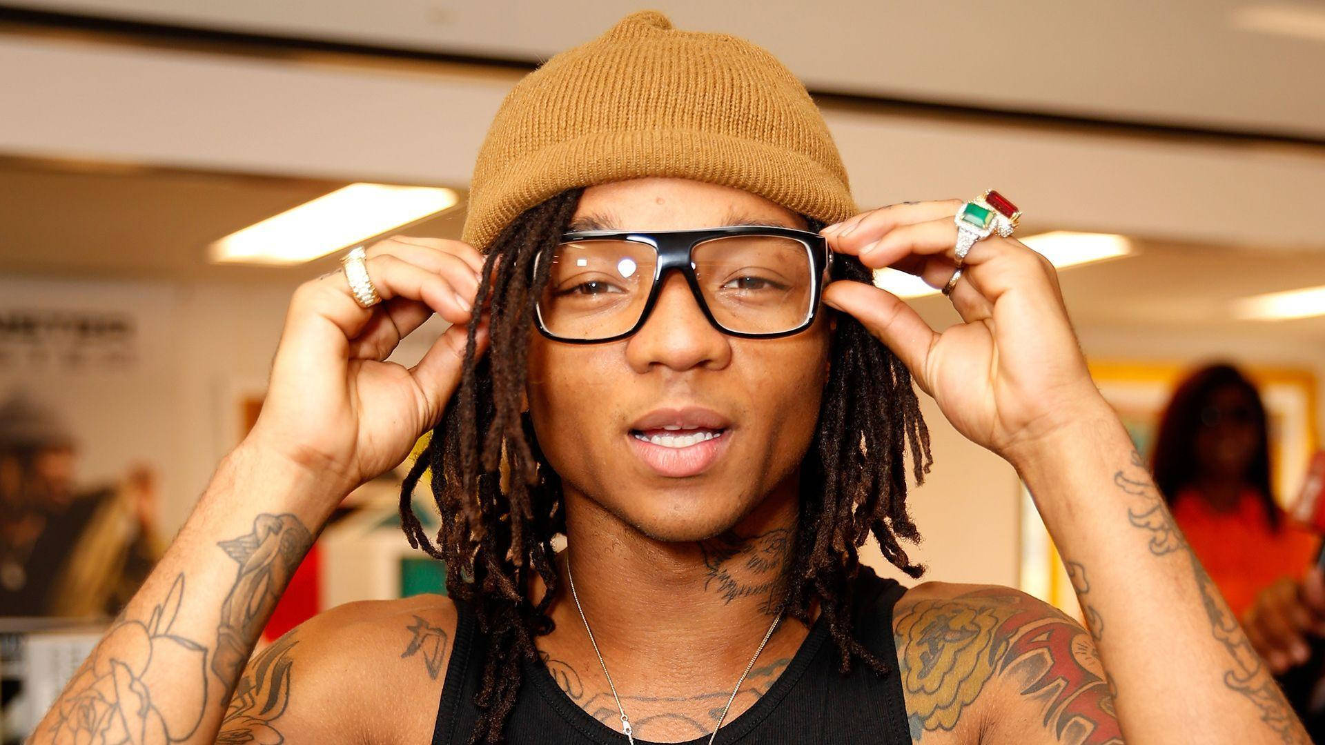 Swae Lee 桌布 壁紙 — 一名留著辮子頭和刺青的男子在室內戴著棕褐色無簷便帽，調整超大黑色眼鏡。