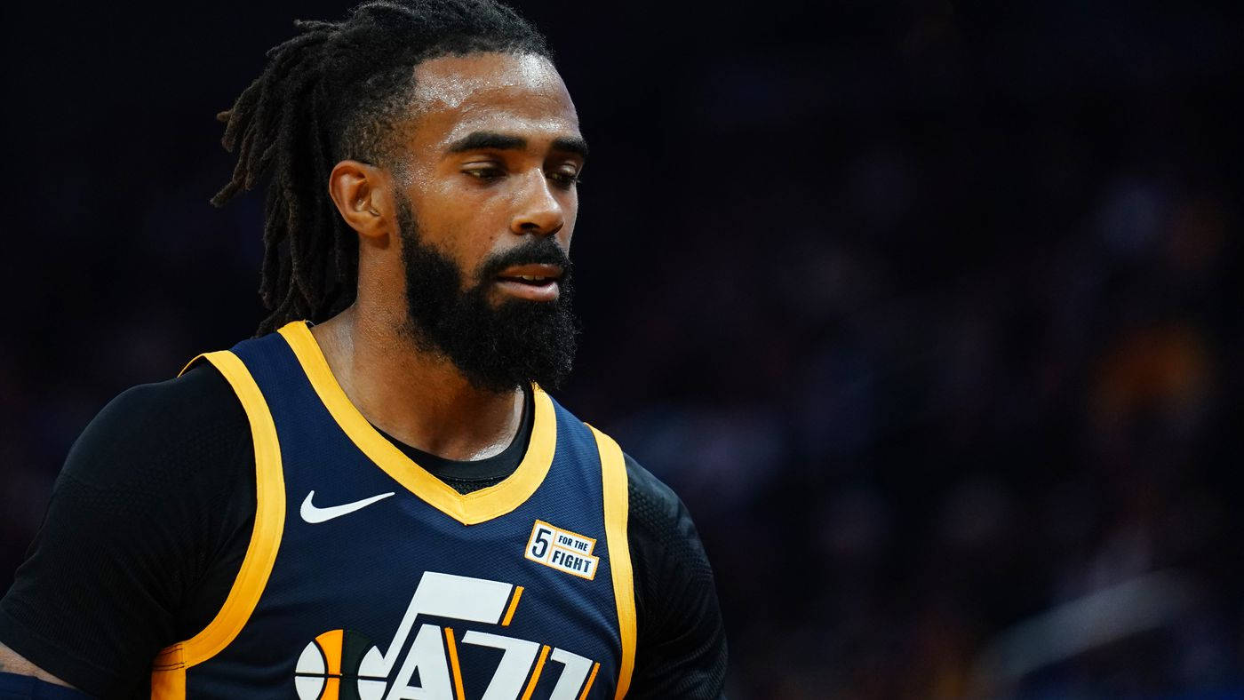 mike conley 桌布 壁紙 — 一名留著辮子頭和鬍鬚、專注的籃球運動員，在場館燈光下穿著海軍藍黃色的爵士隊球衣。