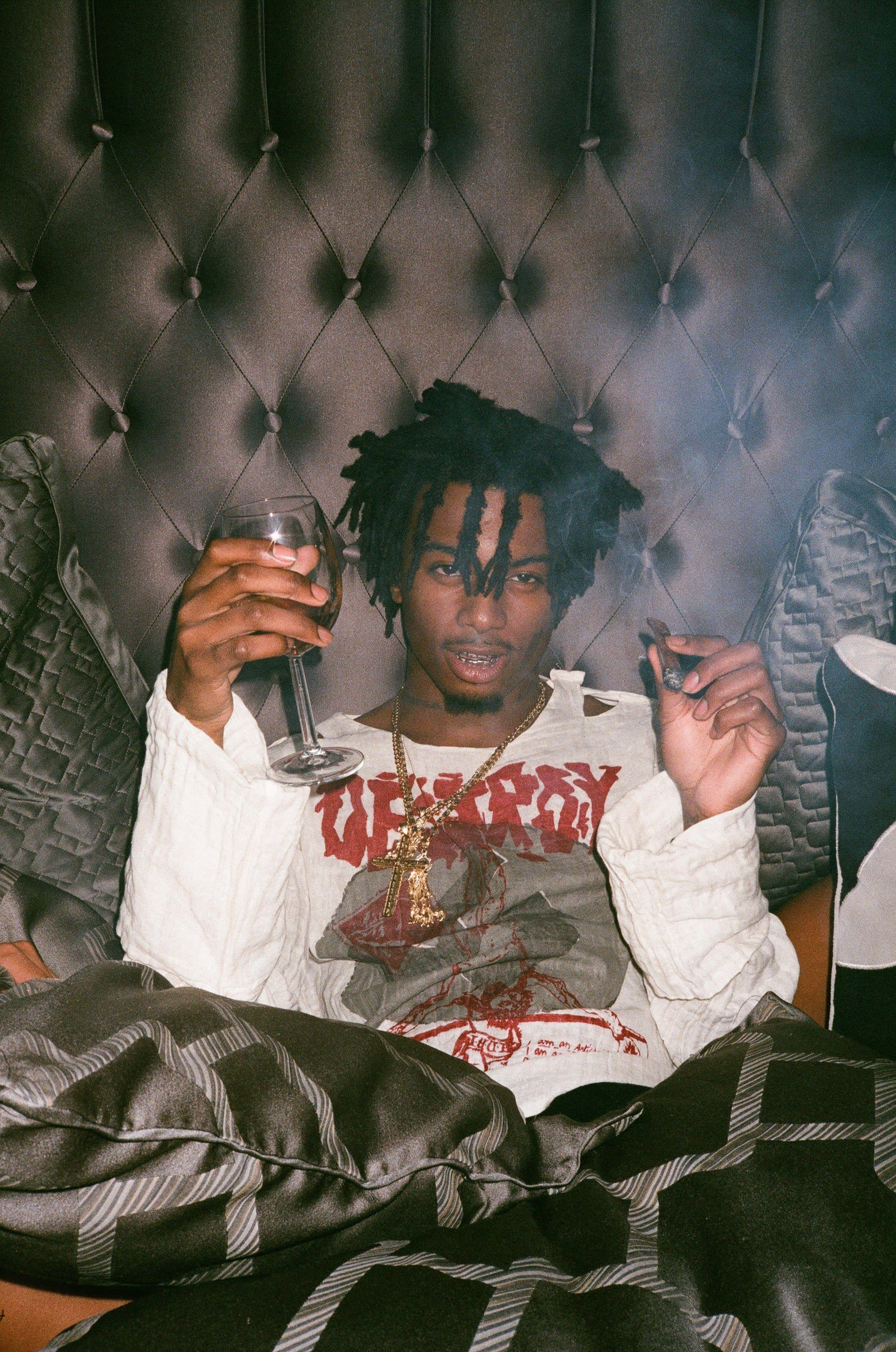 Playboi Carti iPhone 桌布 壁紙 — 一名留著辮子頭的男人在昏暗的房間裡，靠在一張簇絨皮革沙發上，手持酒杯和香煙。