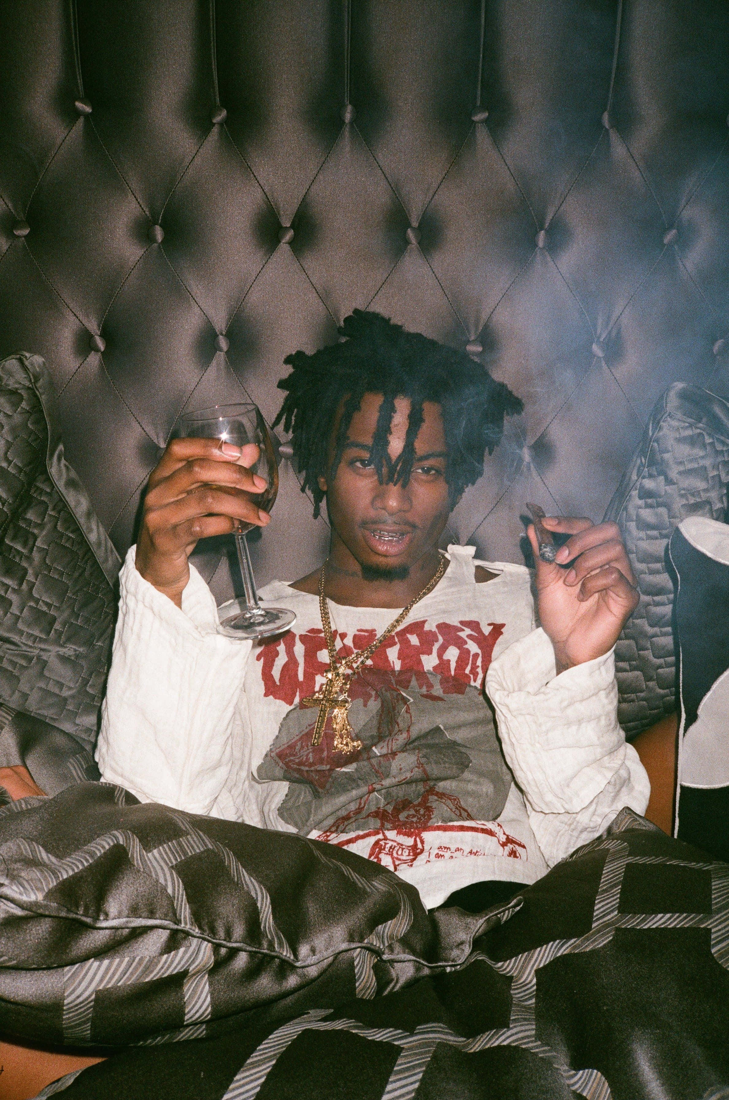 Playboi Carti HD 桌布 壁紙 — 一名留著辮子頭的男人在簇絨皮革沙發上放鬆，手持酒杯並在昏暗燈光下抽煙。