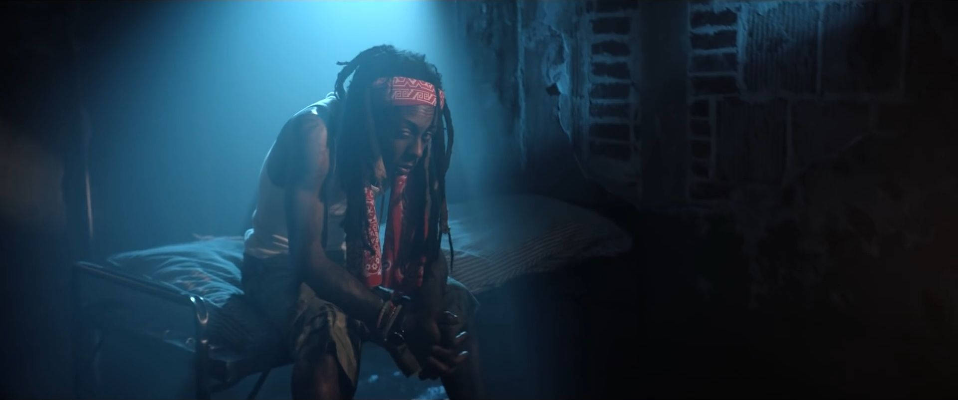Lil Wayne 桌布 壁紙 — 一名留著辮子頭的男人蜷縮著坐在床上，房間光線昏暗，藍色燈光，牆壁剝落，百葉窗破舊。