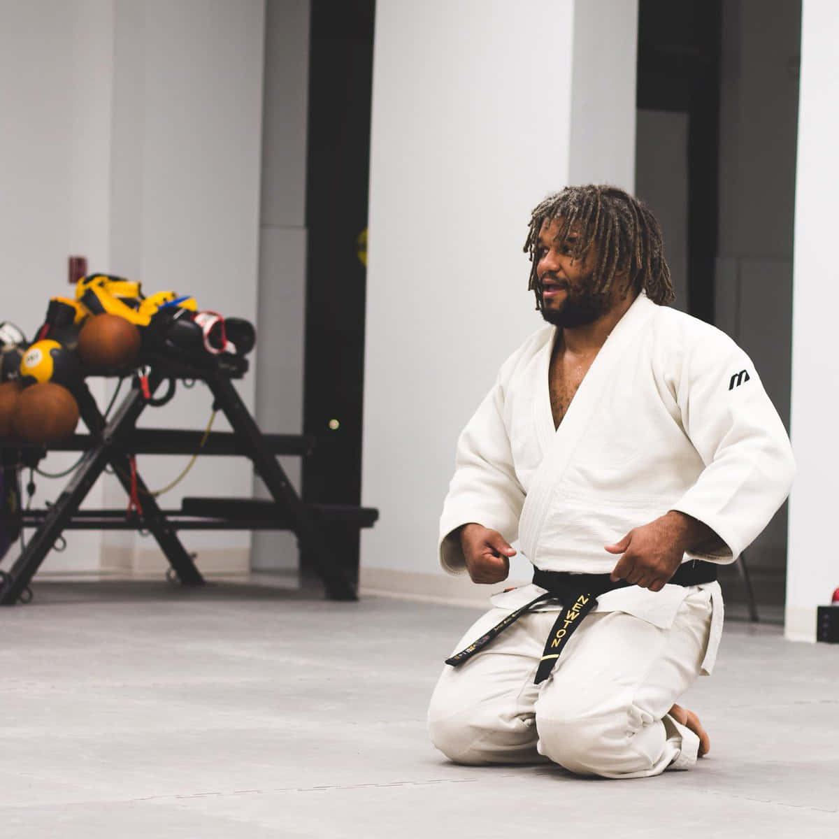 martial arts 桌布 壁紙 — 一名留著辮子頭的男子，穿著白色柔道服和黑色腰帶，跪在現代訓練健身房中。