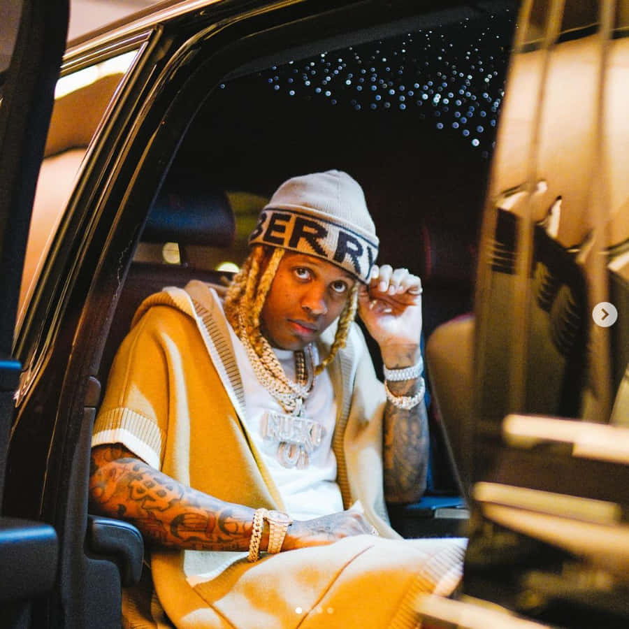 Lil Durk 桌布 壁紙 — 一名留著辮子頭髮、有刺青的男子，戴著毛帽和金鍊，坐在豪華車內，直視鏡頭。