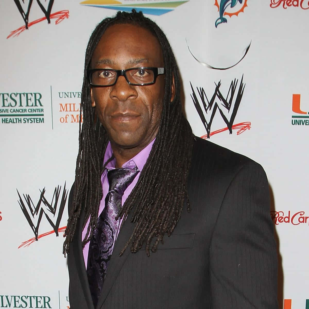 WWE 桌布 壁紙 — 一名留著長辮子、戴眼鏡的男子，身穿黑色西裝和紫色領帶，背景是品牌活動場景。