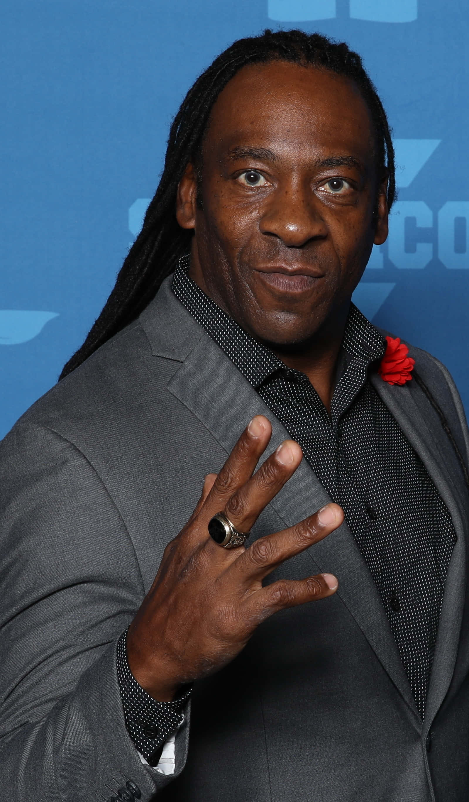 WWE 桌布 壁紙 — 一名留著長辮子、身穿灰色西裝的男子在藍色背景前微笑並做出手勢。