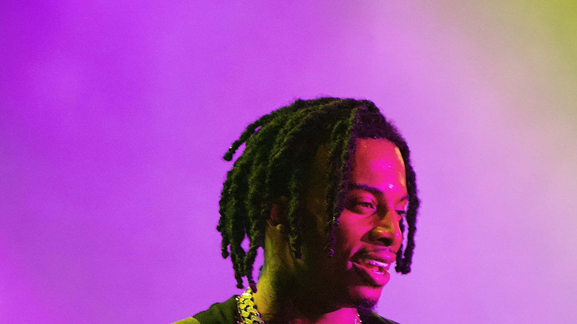 Playboi Carti 桌布 壁紙 — 一名留著長辮子的男人在充滿活力的紫色和黃色舞台燈光下微笑。