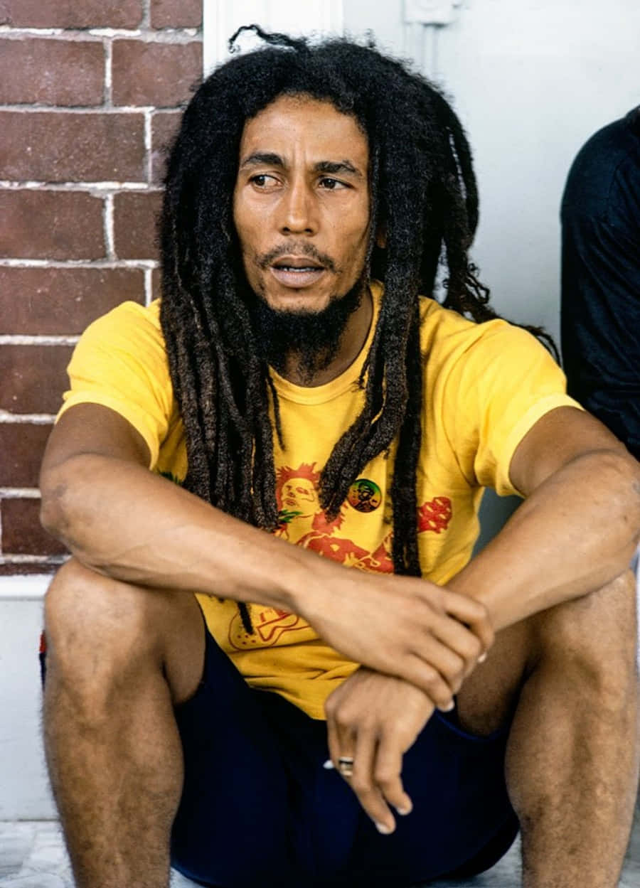 Bob Marley 桌布 壁紙 — 一名留著長髮辮的男人盤腿坐在磚牆前，穿著鮮黃色T恤。