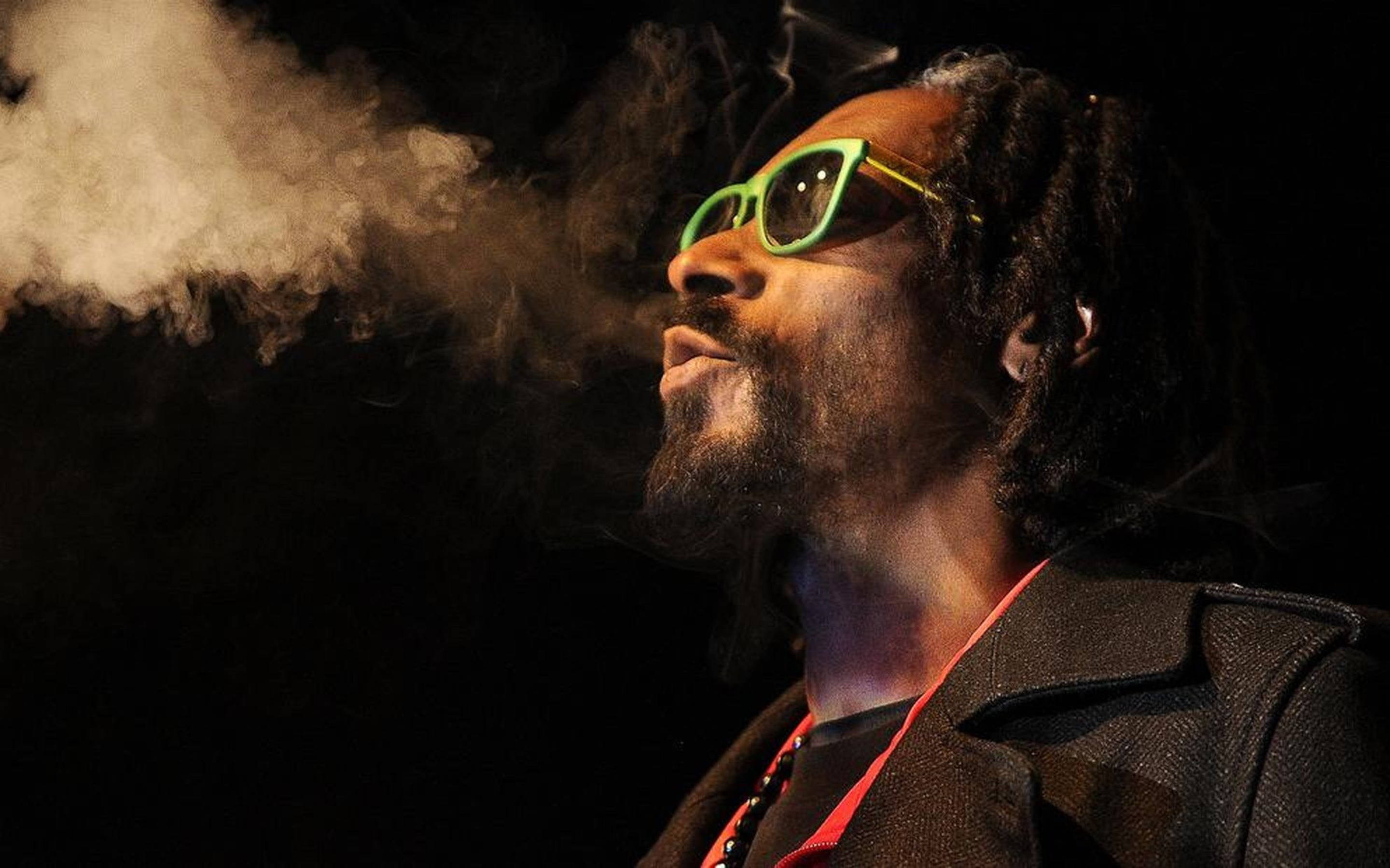 Snoop Dogg 桌布 壁紙 — 一名留著雷鬼辮、戴著亮綠色眼鏡的男子，在深色背景前呼出一團濃煙。