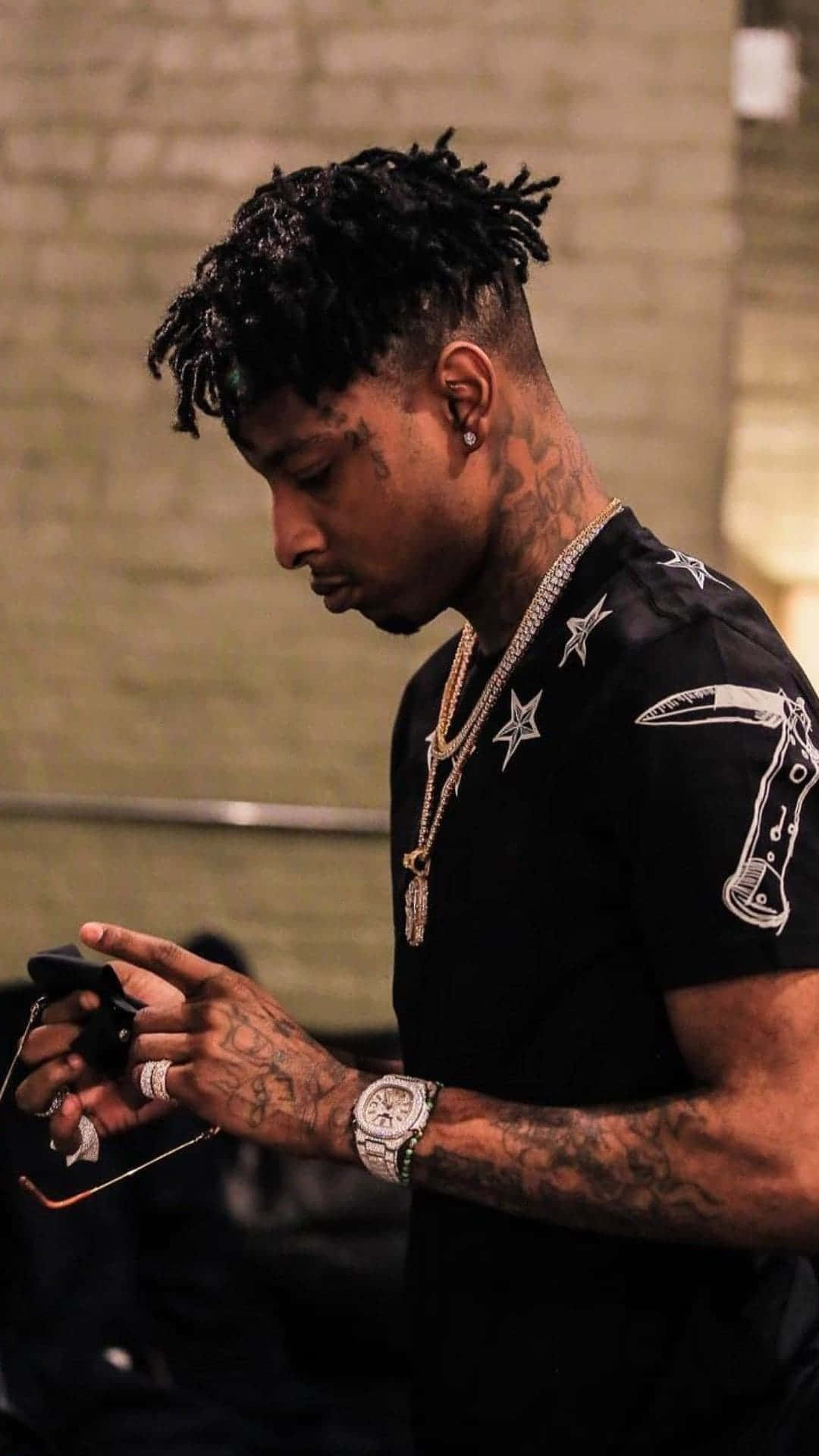 21 Savage 桌布 壁紙 — 一名留著雷鬼辮、有刺青的男子，穿著黑色星星印花襯衫和鑽石鏈條，低頭看著手機。