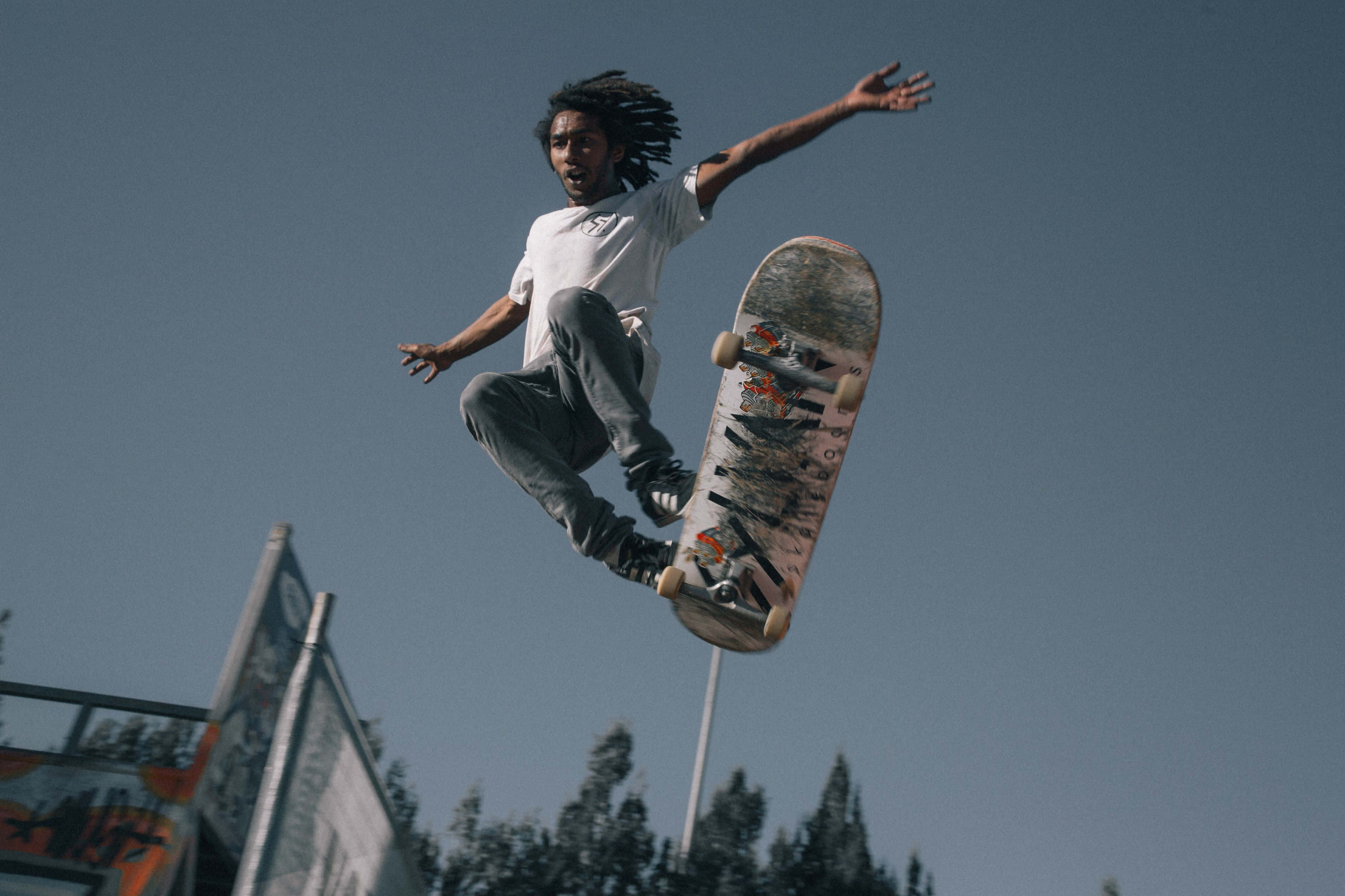 Skater Boy 桌布 壁紙 — 一名留著雷鬼辮的滑板手在藍天白雲下，於背景有樹木的滑板公園上空飛翔。
