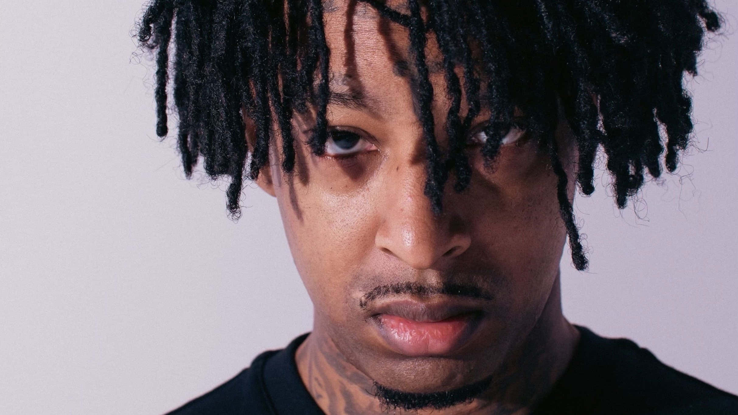 21 Savage 桌布 壁紙 — 一名留著黑色辮子、身上有刺青的男子，在純淺灰色背景前凝視的特寫肖像。