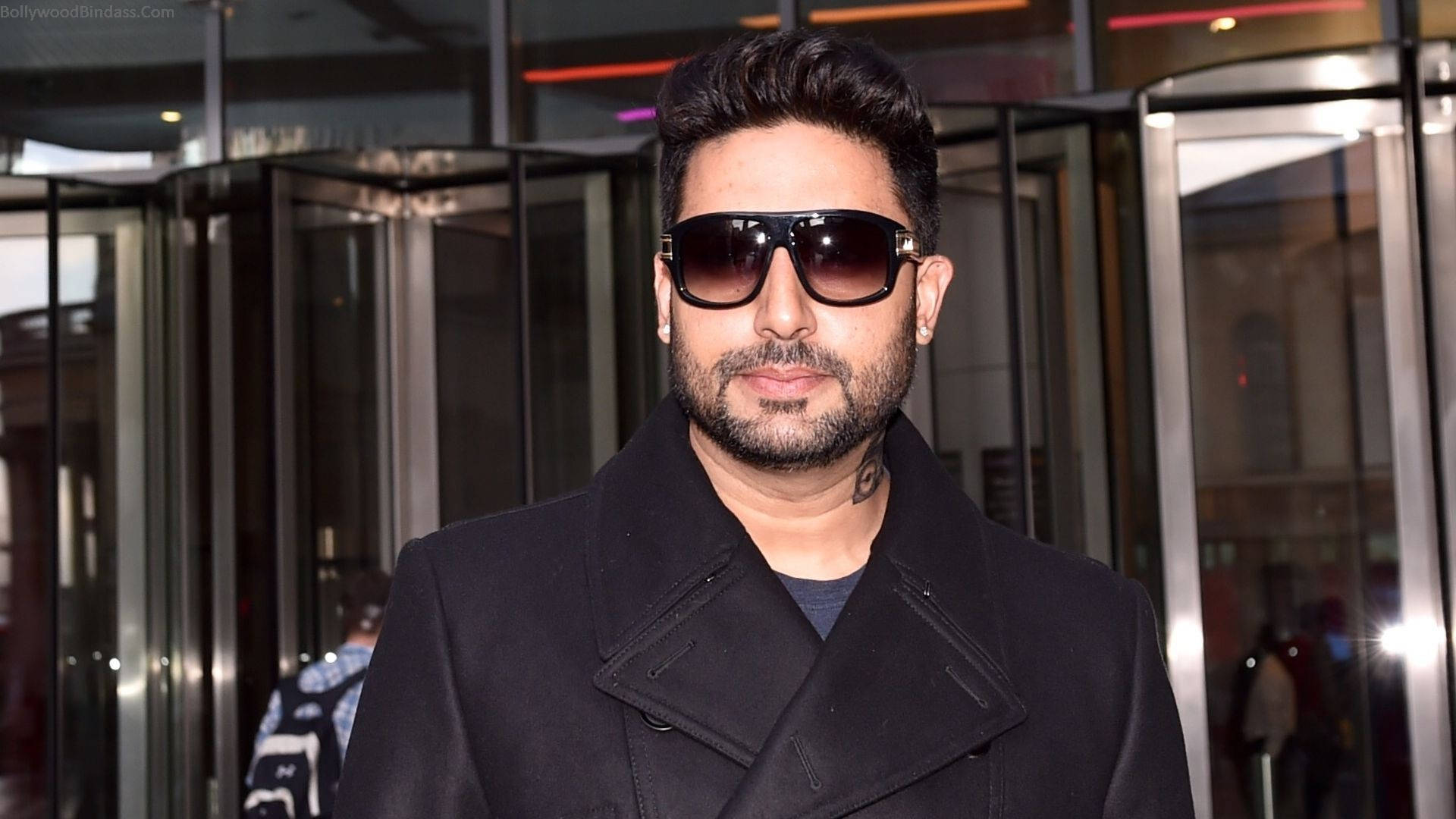 abhishek bachchan 桌布 壁紙 — 一名留鬍子、戴太陽眼鏡的男子在夜晚自信地站在玻璃和金屬旋轉門前。