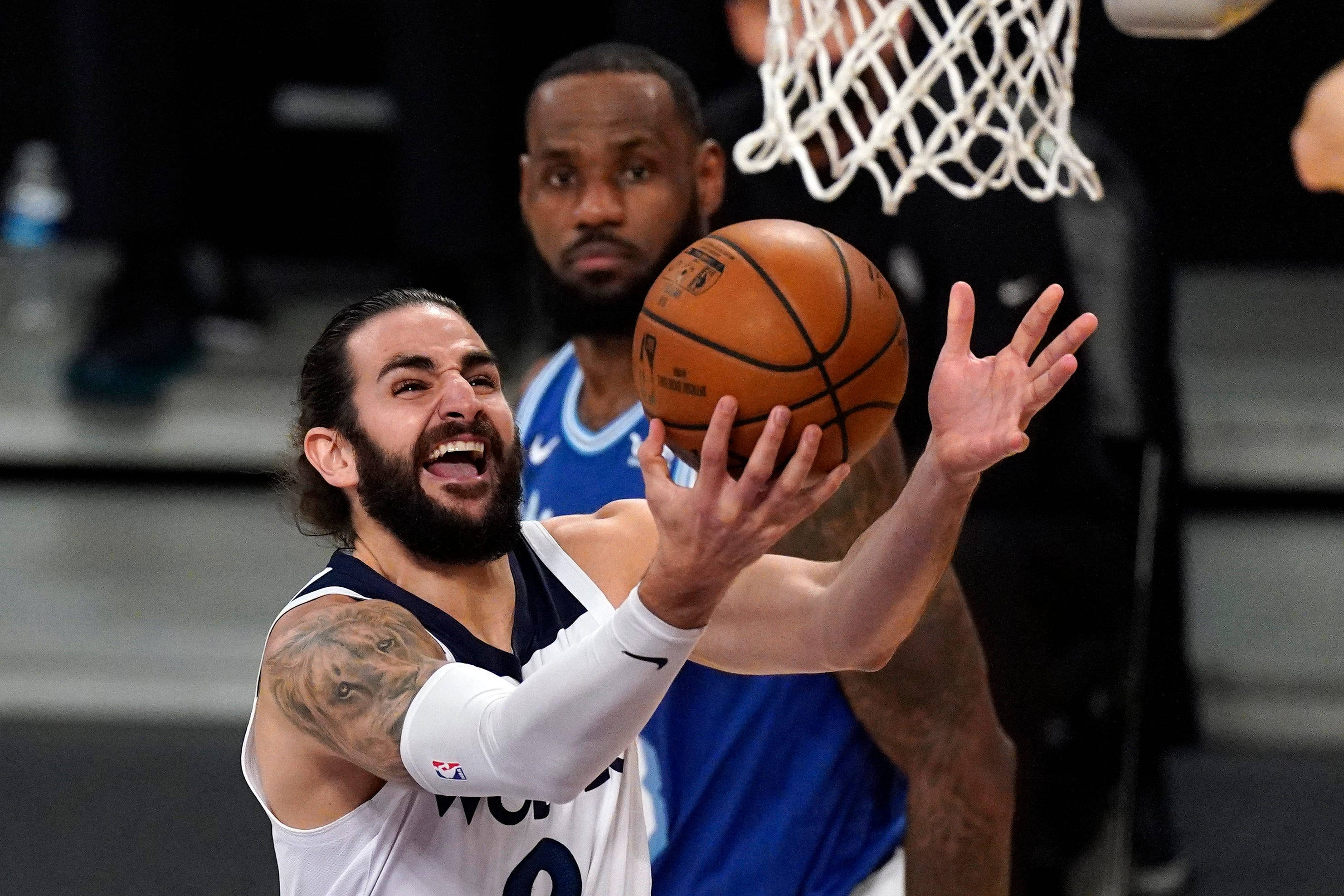 Ricky Rubio 桌布 壁紙 — 一名留鬍子、手臂有刺青的籃球運動員跳起投籃，防守球員在場館燈光下緊張注視。