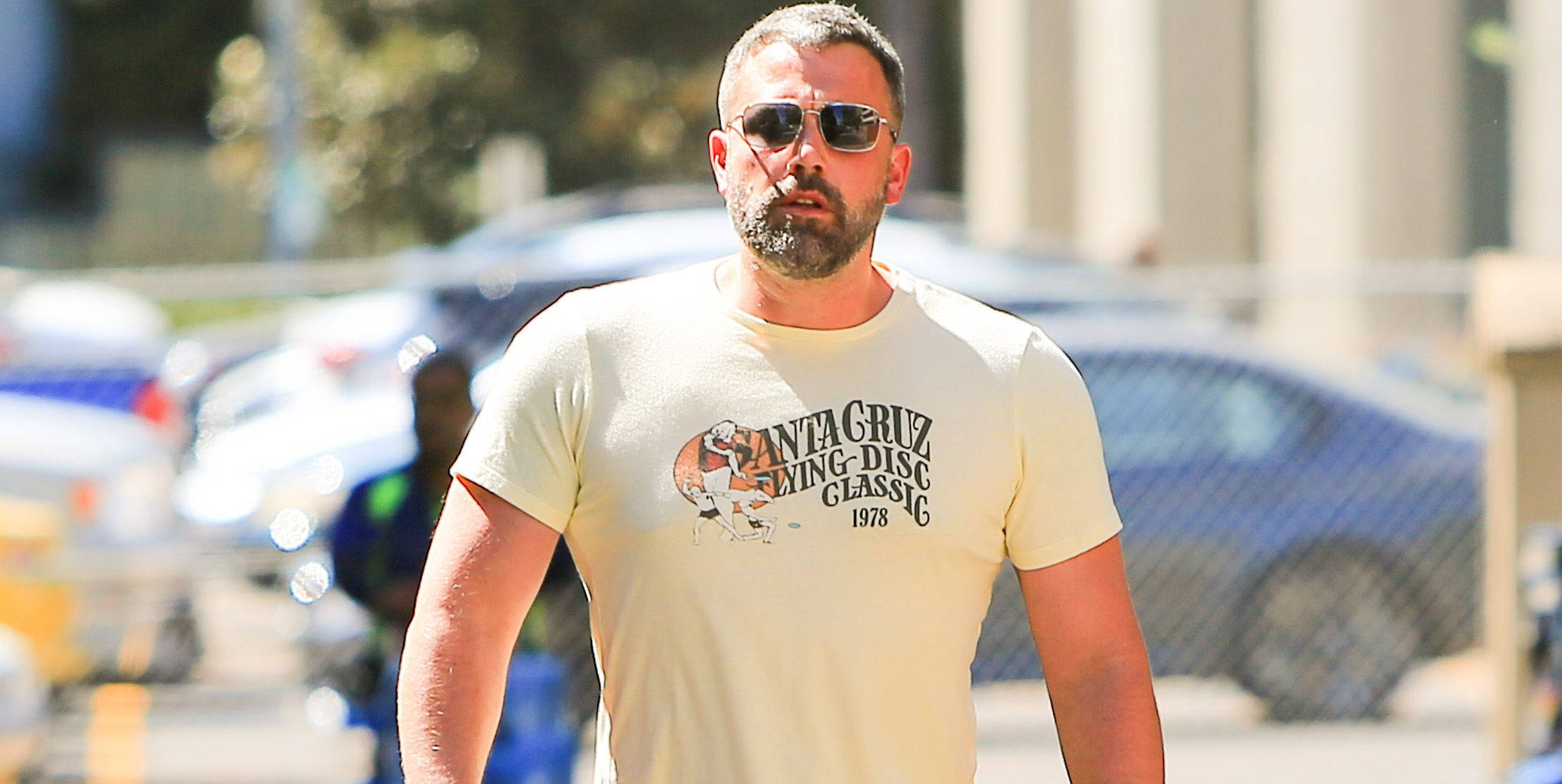 Ben Affleck 桌布 壁紙 — 一名留鬍鬚、戴太陽眼鏡的男子在明亮的陽光下穿著復古Santa Cruz T恤在戶外行走。