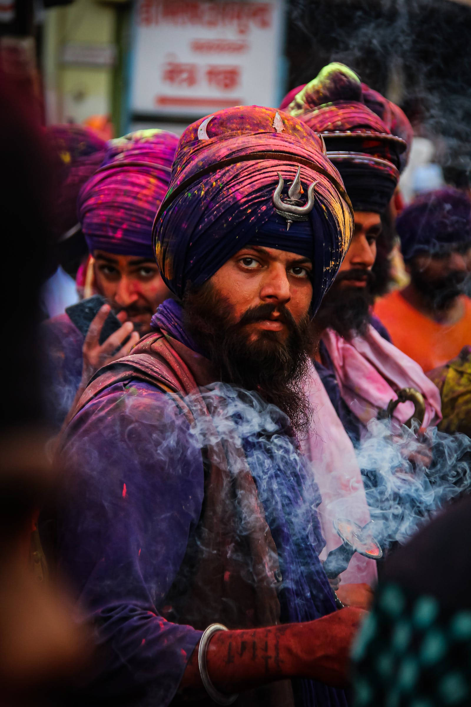 Holi 桌布 壁紙 — 一名留鬍鬚、戴彩色頭巾的男子，在充滿粉末色彩的熱鬧灑紅節人群中吸煙。
