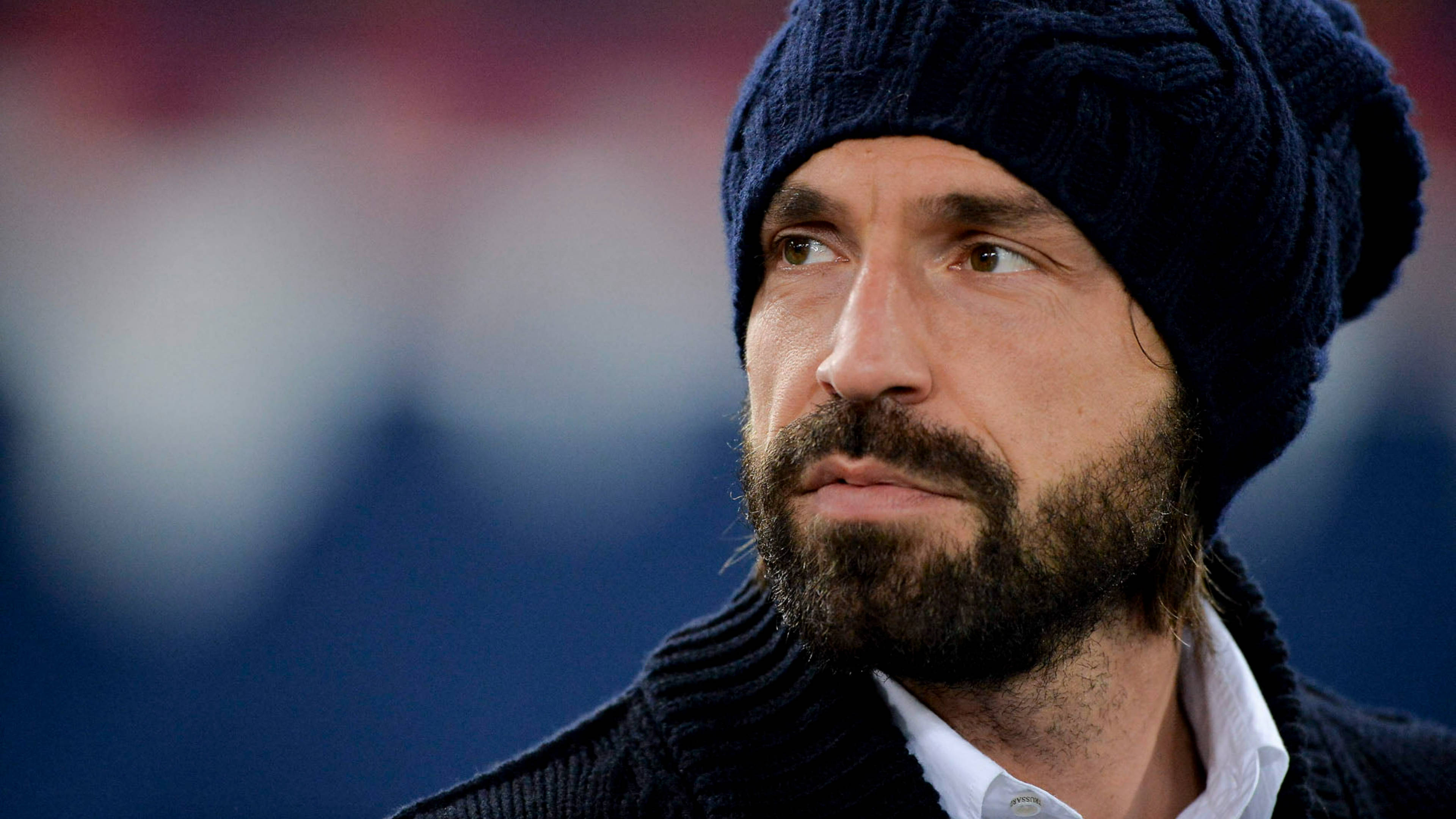 andrea pirlo 桌布 壁紙 — 一名留鬍鬚、戴深色針織毛帽的男子，在模糊的體育場背景下若有所思地望向一旁。