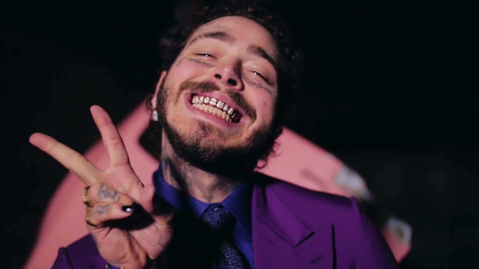 Post Malone 桌布 壁紙 — 一名留鬍鬚、戴鑽石牙套的男子在深色背景下穿著紫色西裝，微笑著比出和平手勢。