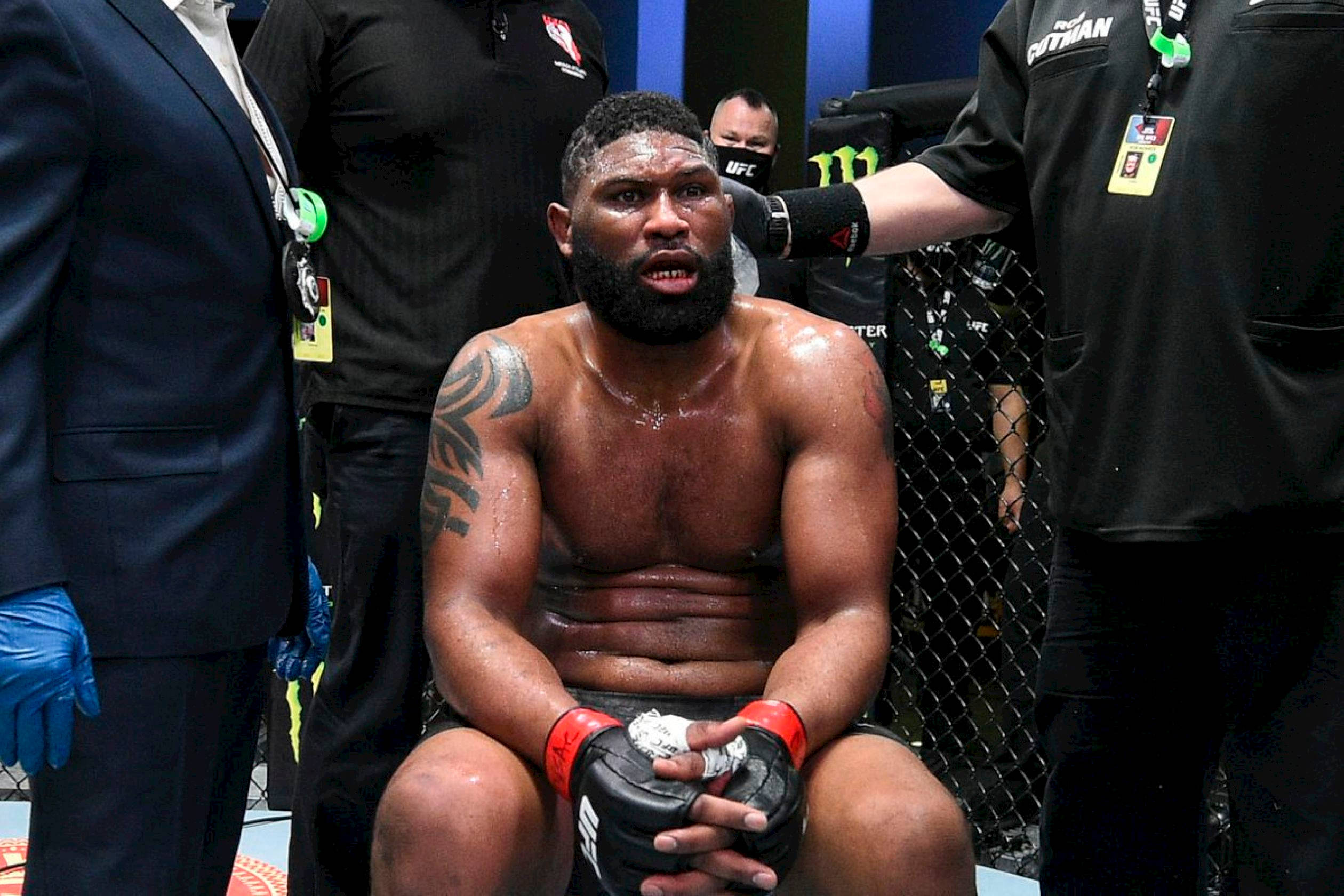 UFC 桌布 壁紙 — 一名留鬍鬚、滿身大汗的肌肉發達拳手，在殘酷比賽後筋疲力竭地坐在八角籠中，周圍是工作人員。