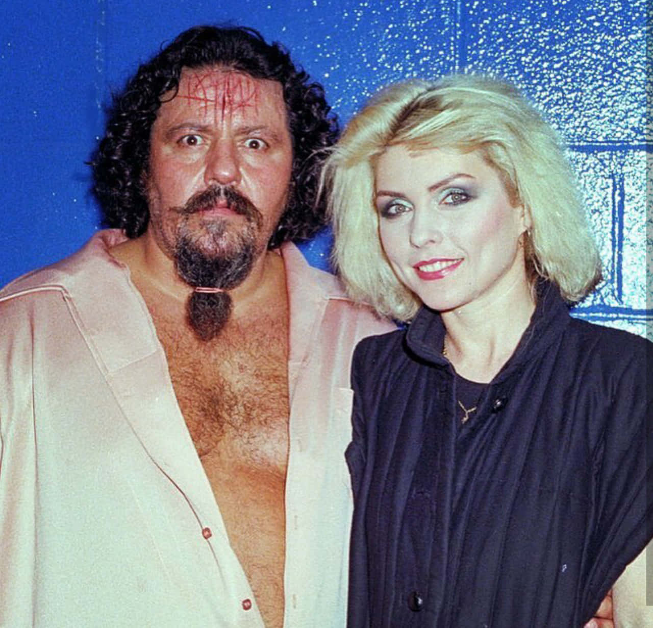 Lou Albano 桌布 壁紙 — 一名留鬍鬚、臉上塗有紅色顏料的男子，站在一位身穿深色襯衫的金髮女子旁邊，背景是閃爍的藍色。