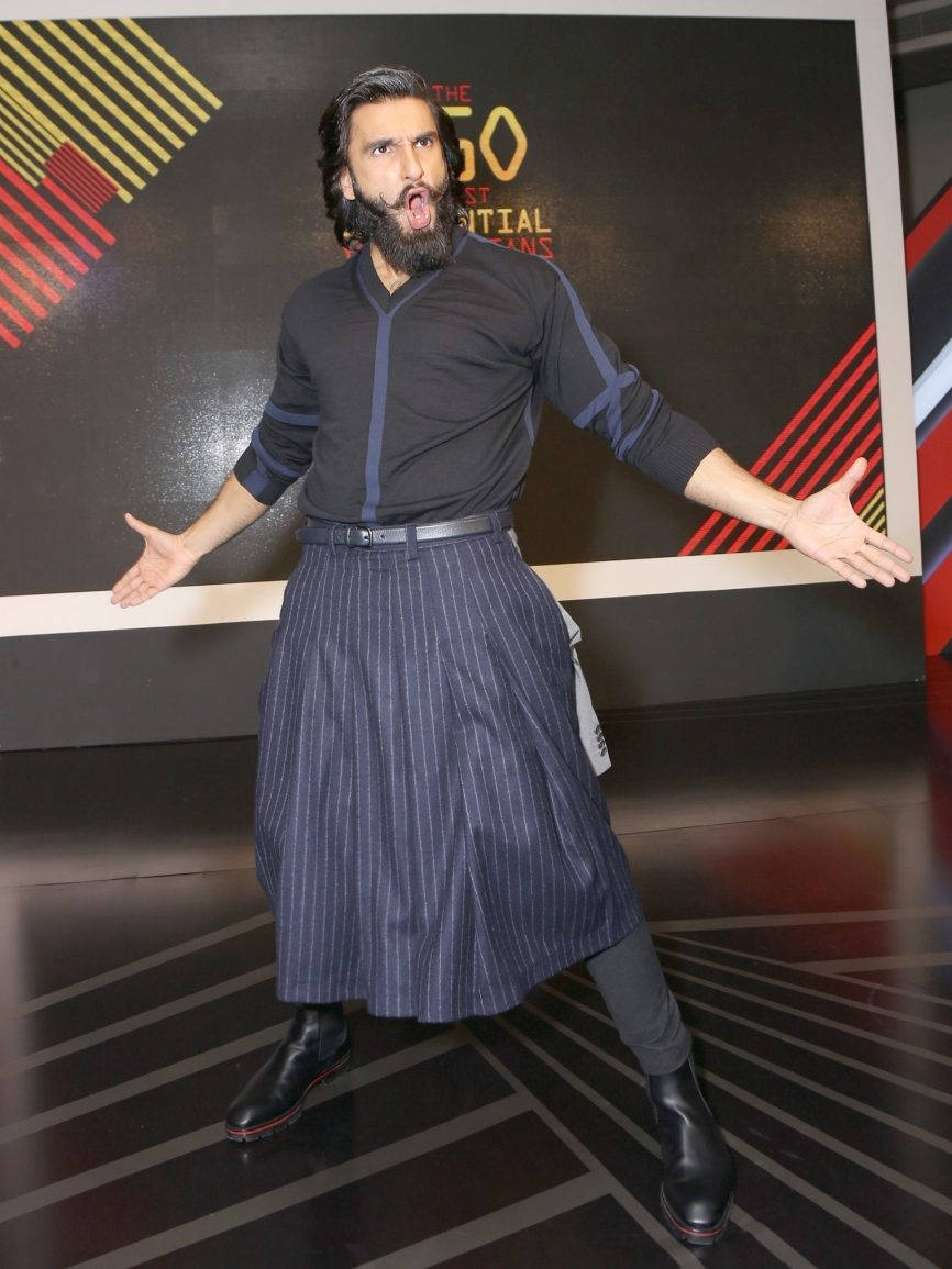 Ranveer Singh 桌布 壁紙 — 一名留鬍鬚的男子穿著深色細條紋裙子，在幾何圖案背景前張開雙臂擺出俏皮的姿勢。