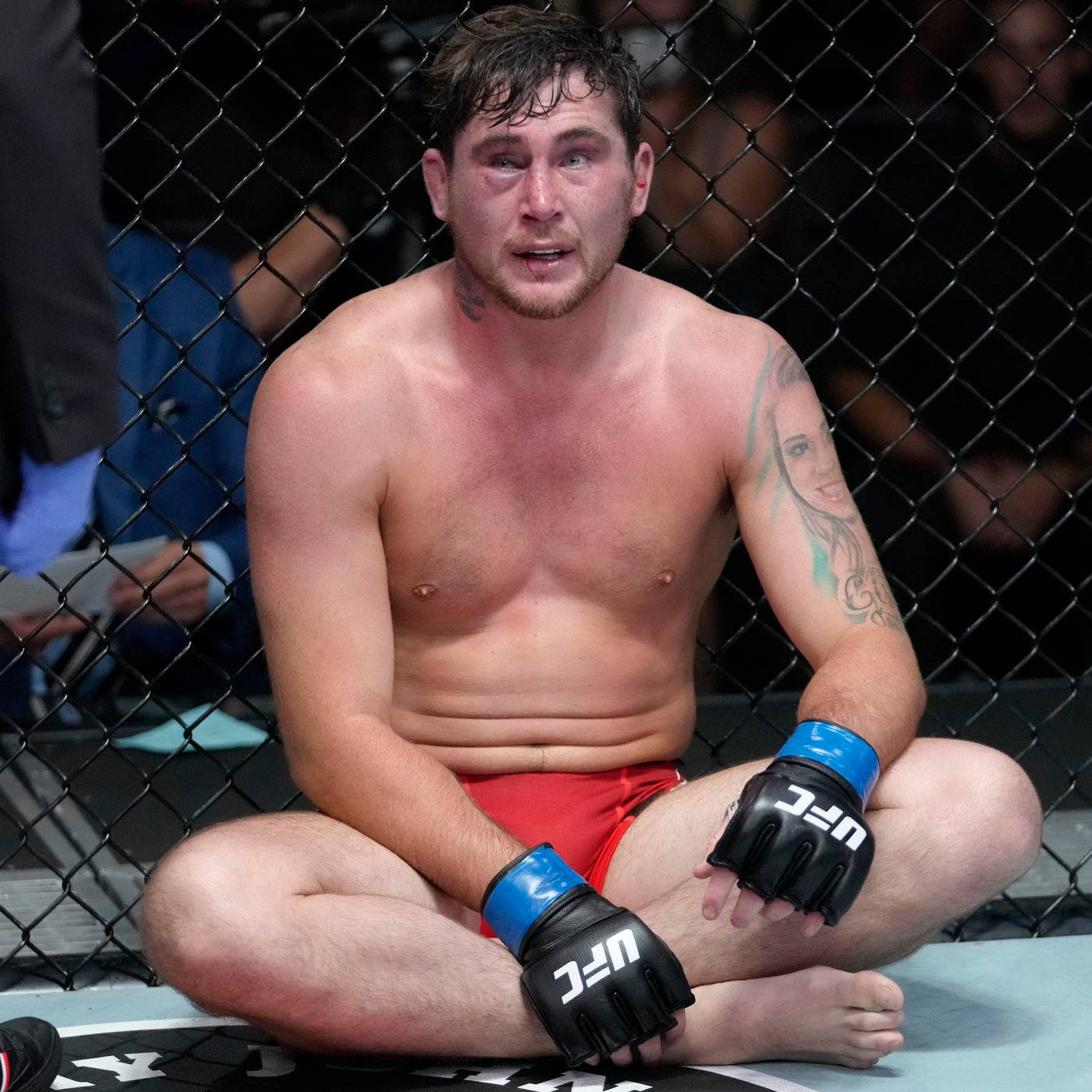 UFC 桌布 壁紙 — 一名疲憊的UFC拳手在殘酷比賽後，滿臉鮮血、手臂有刺青，坐在八角籠地板上。