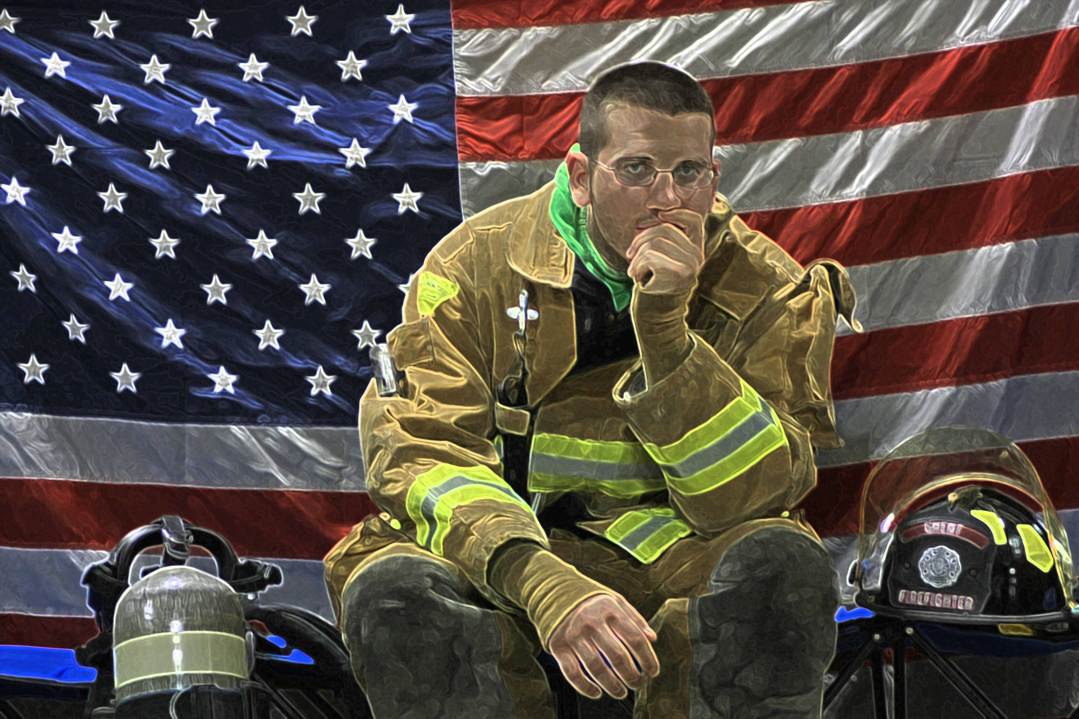 Firefighter 桌布 壁紙 — 一名疲憊的消防員全副裝備，頭盔和氧氣罐放在身旁，若有所思地坐在一面大型美國國旗前。