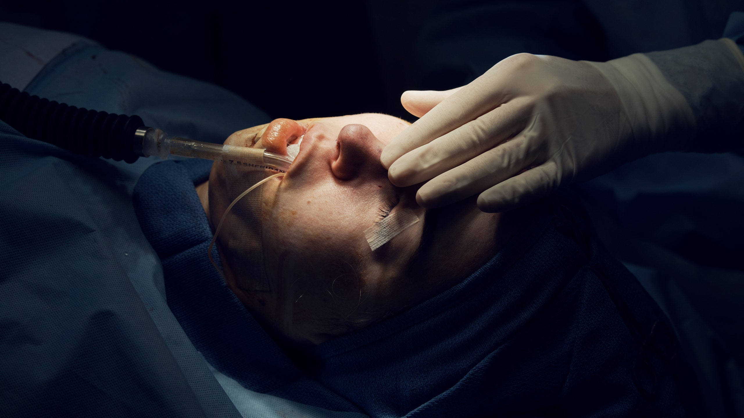 Medical 桌布 壁紙 — 一名病人昏迷不醒地躺在手術台上，戴著手套的手在昏暗的手術燈光下調整氣管內管。