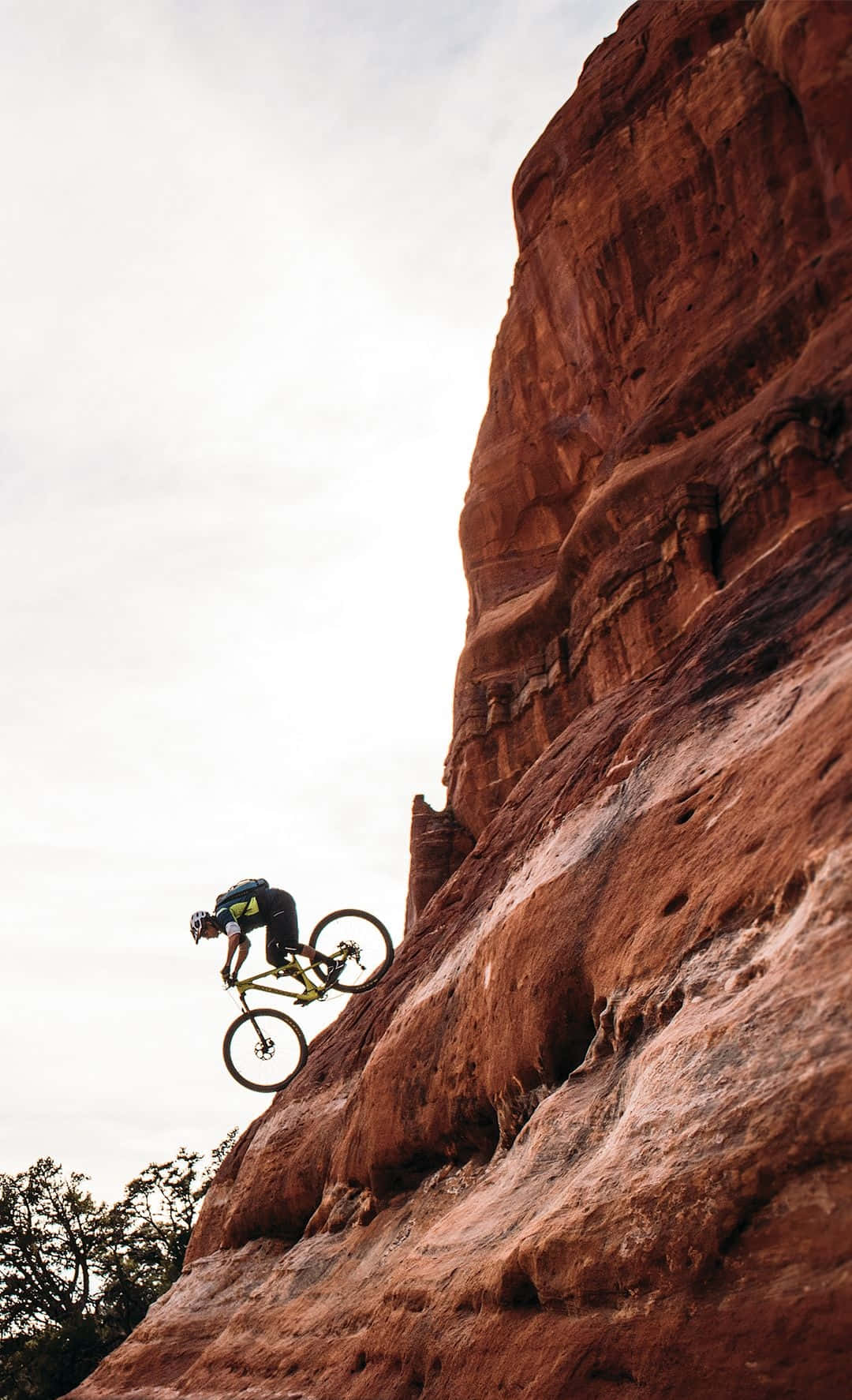 Mountain Bike 桌布 壁紙 — 一名登山自行車手在陰天之下，飛越一座高聳的紅色岩石懸崖。
